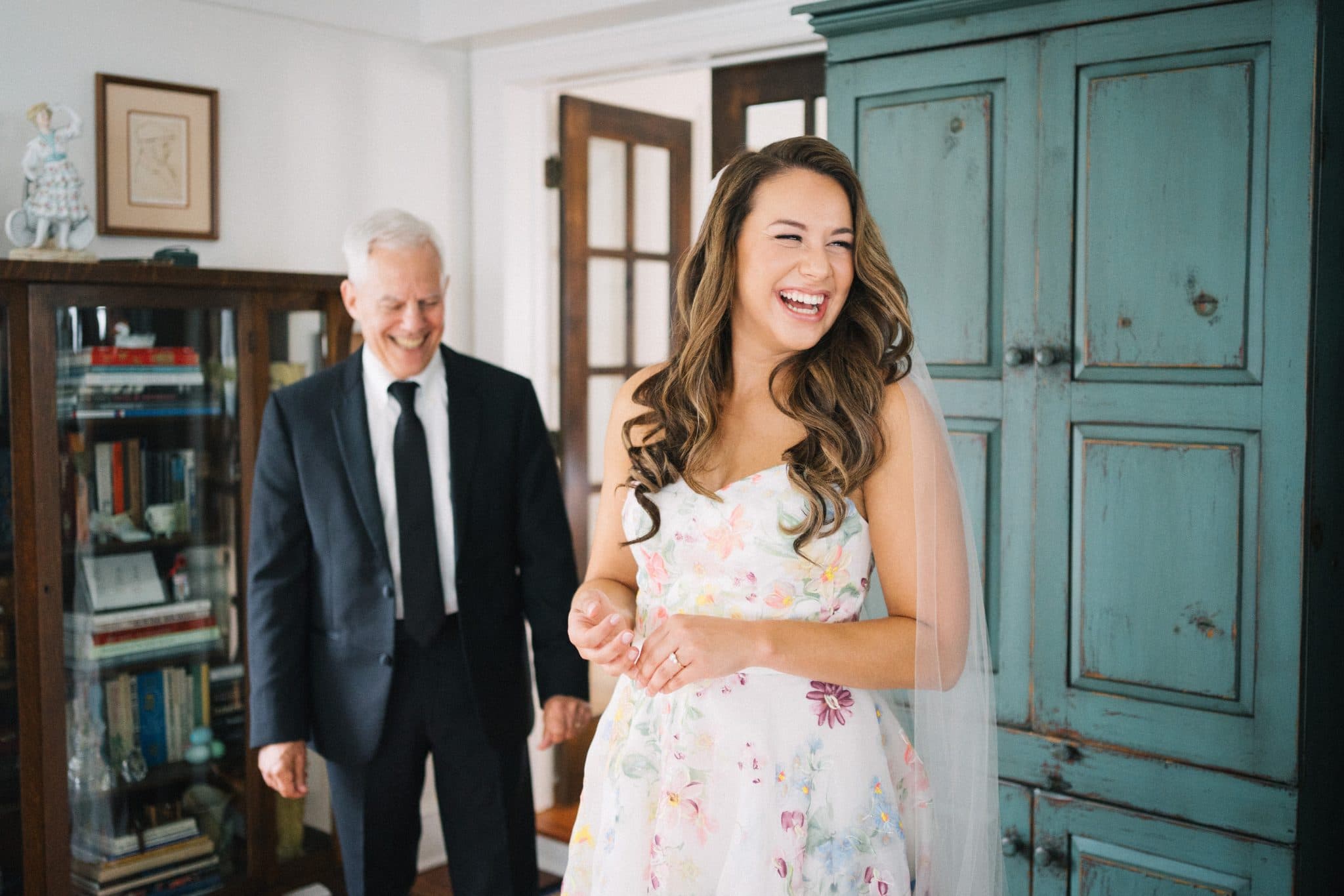 A bride in a floral dress laughs while walking with a man in a suit in a room with a bookshelf and a blue cabinet.