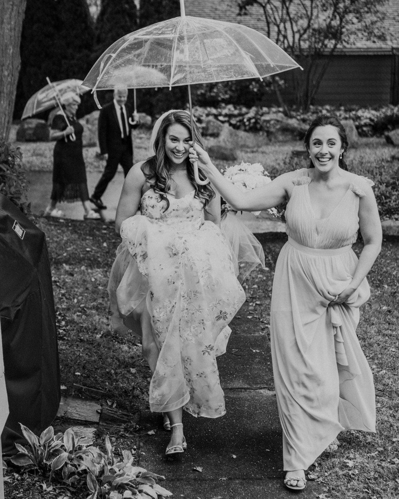 A bride in a floral dress walks under a clear umbrella, accompanied by a woman in a light dress, with another couple in the background.