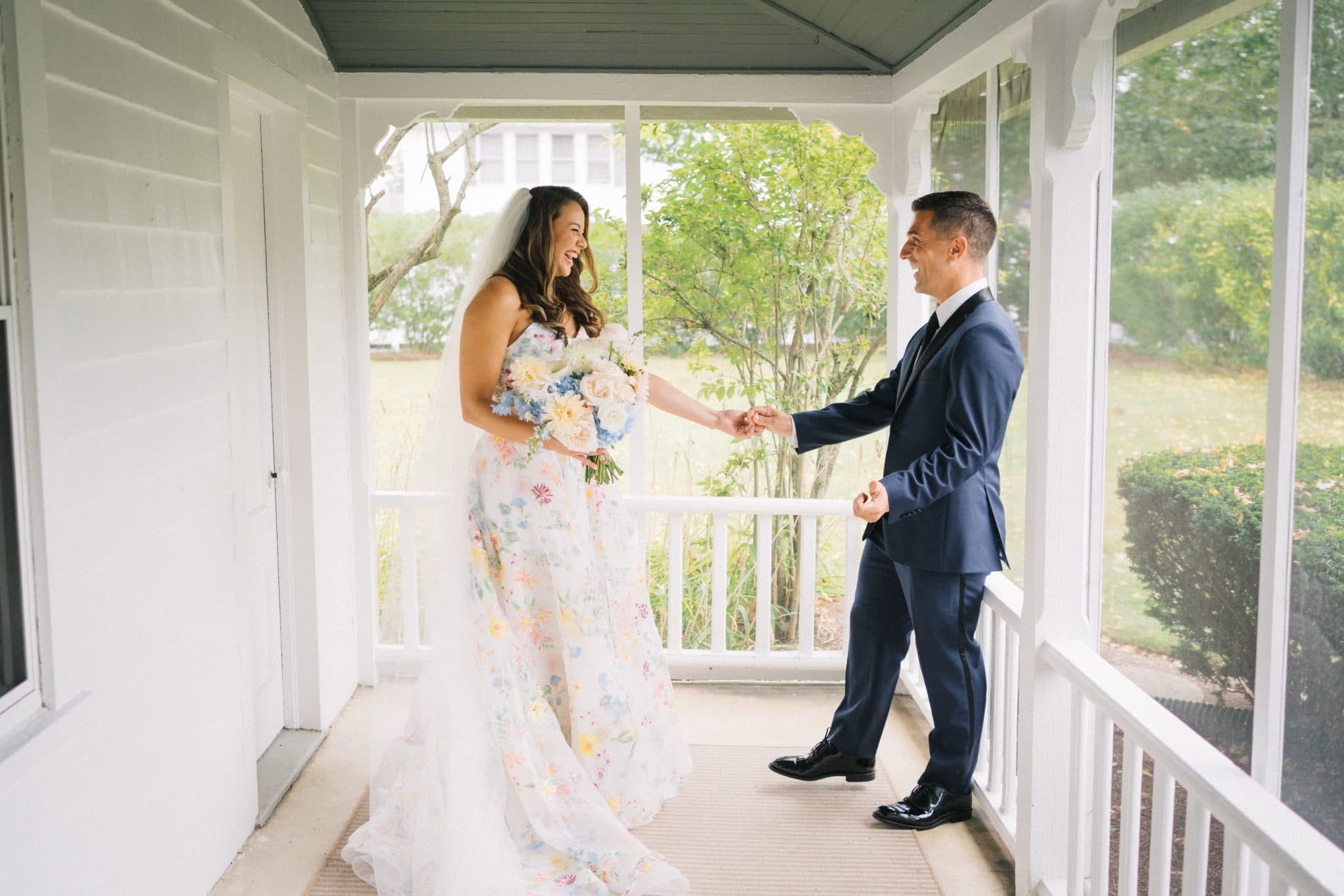 A bride in a floral dress and a groom in a suit hold hands, smiling, on a porch with white railings and a view of greenery outside.