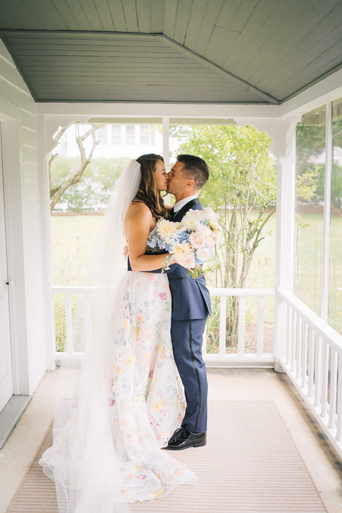 A bride in a floral dress and veil kisses a groom in a blue suit on a porch with white railings and greenery in the background.