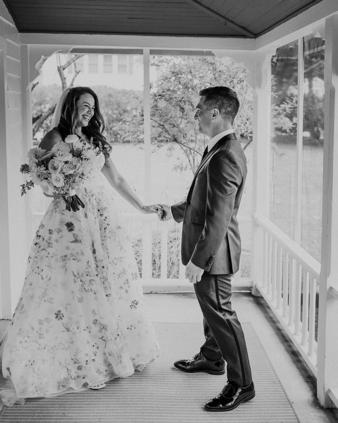 A bride in a floral gown and a groom in a suit hold hands, standing on a porch with glass panels.