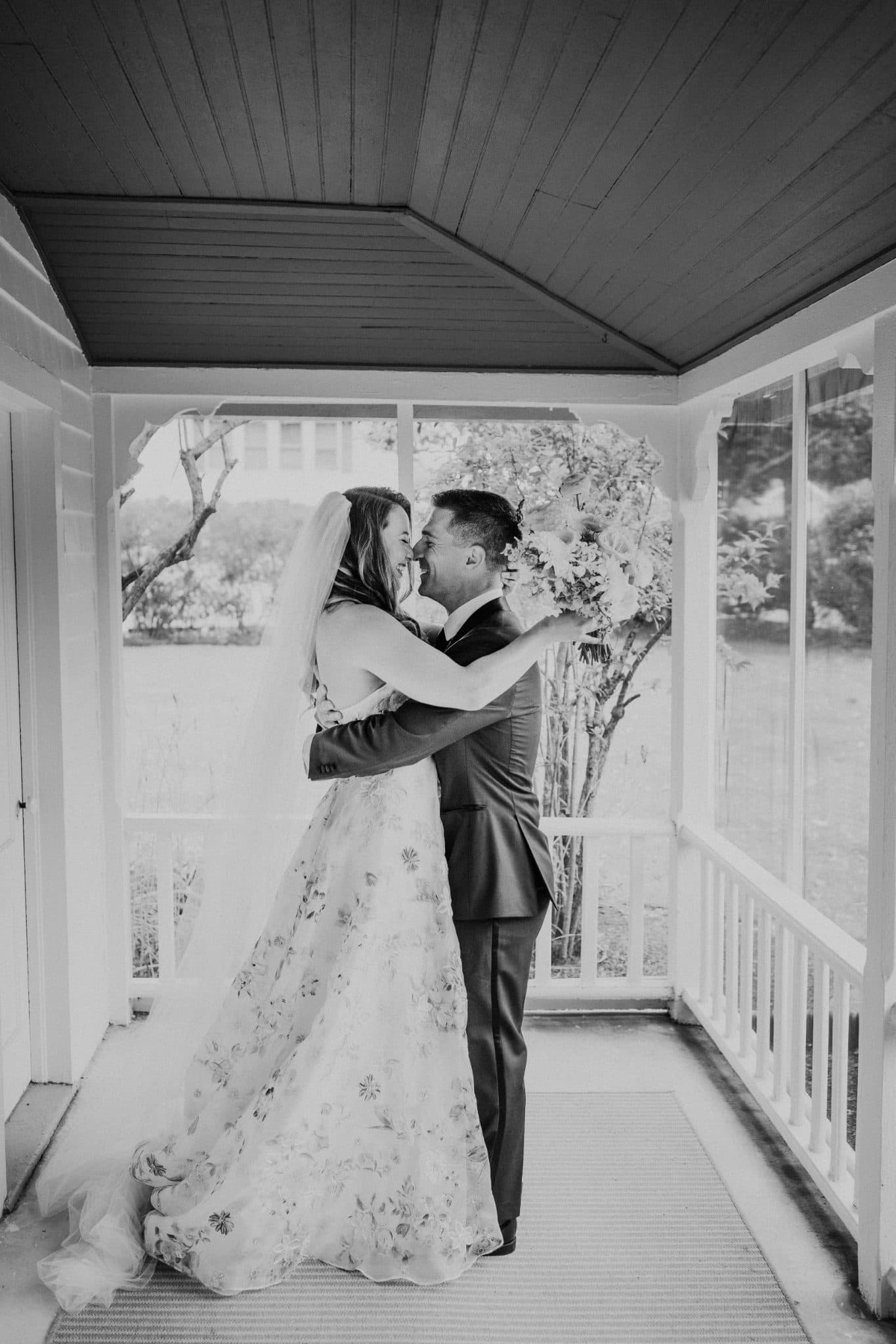 A bride in a floral gown and veil embraces a groom in a suit on a porch with a view of trees and grass.