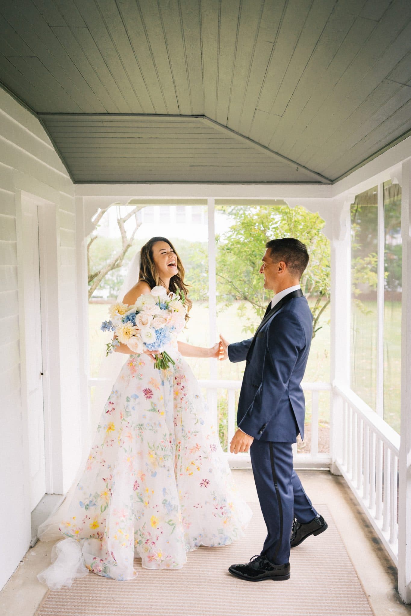 A bride in a floral gown and a groom in a navy suit hold hands on a porch with white railings and a ceiling.