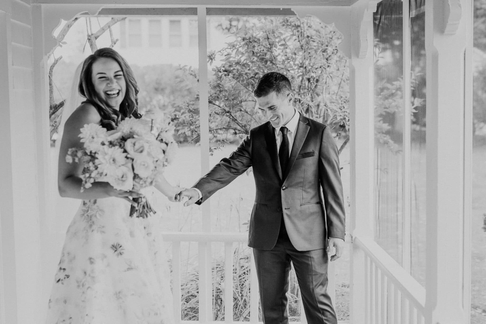A bride in a floral dress and a groom in a suit hold hands, smiling, in a porch setting.