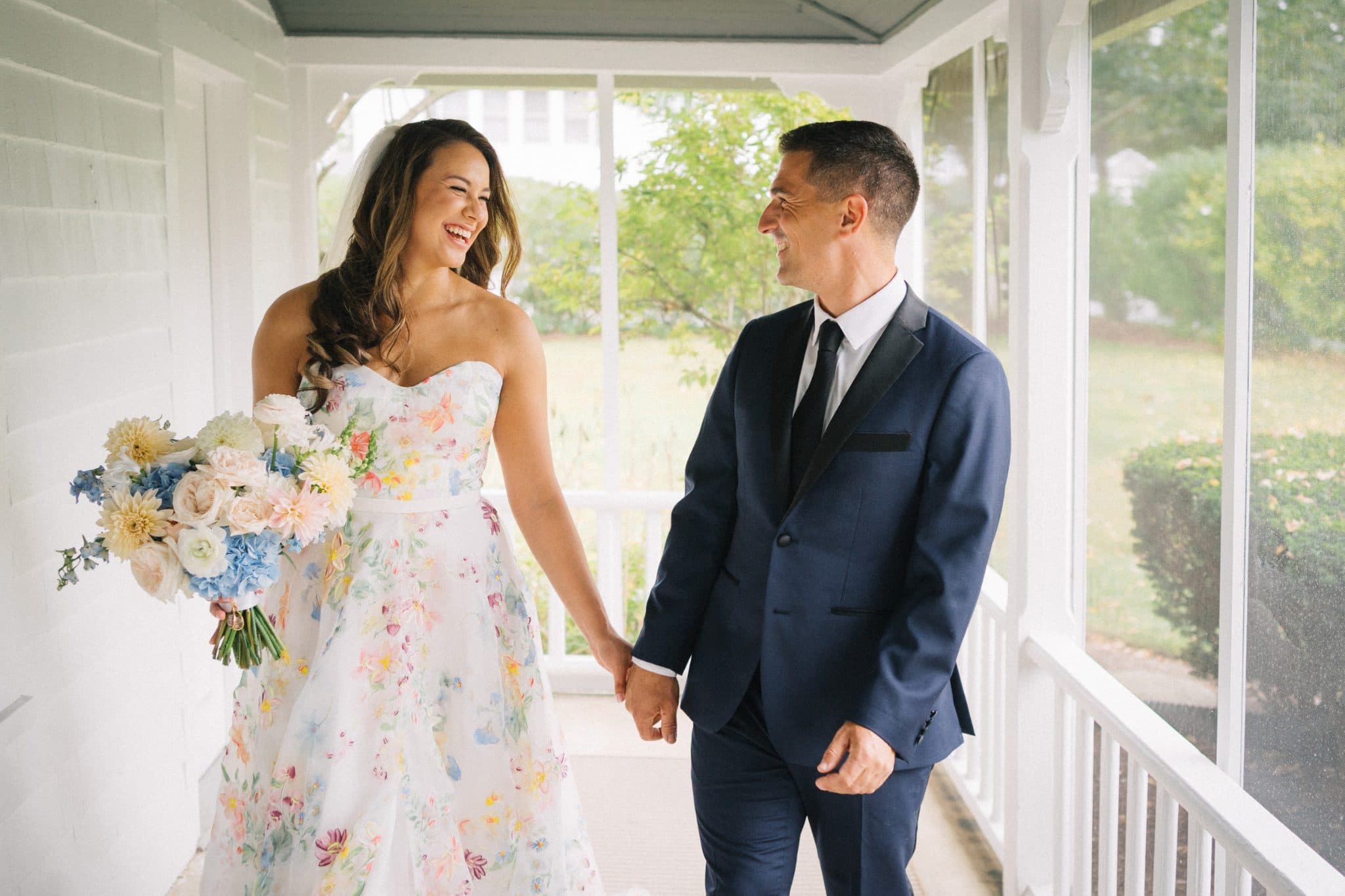 A bride in a floral dress and a groom in a navy suit walk hand in hand on a porch.