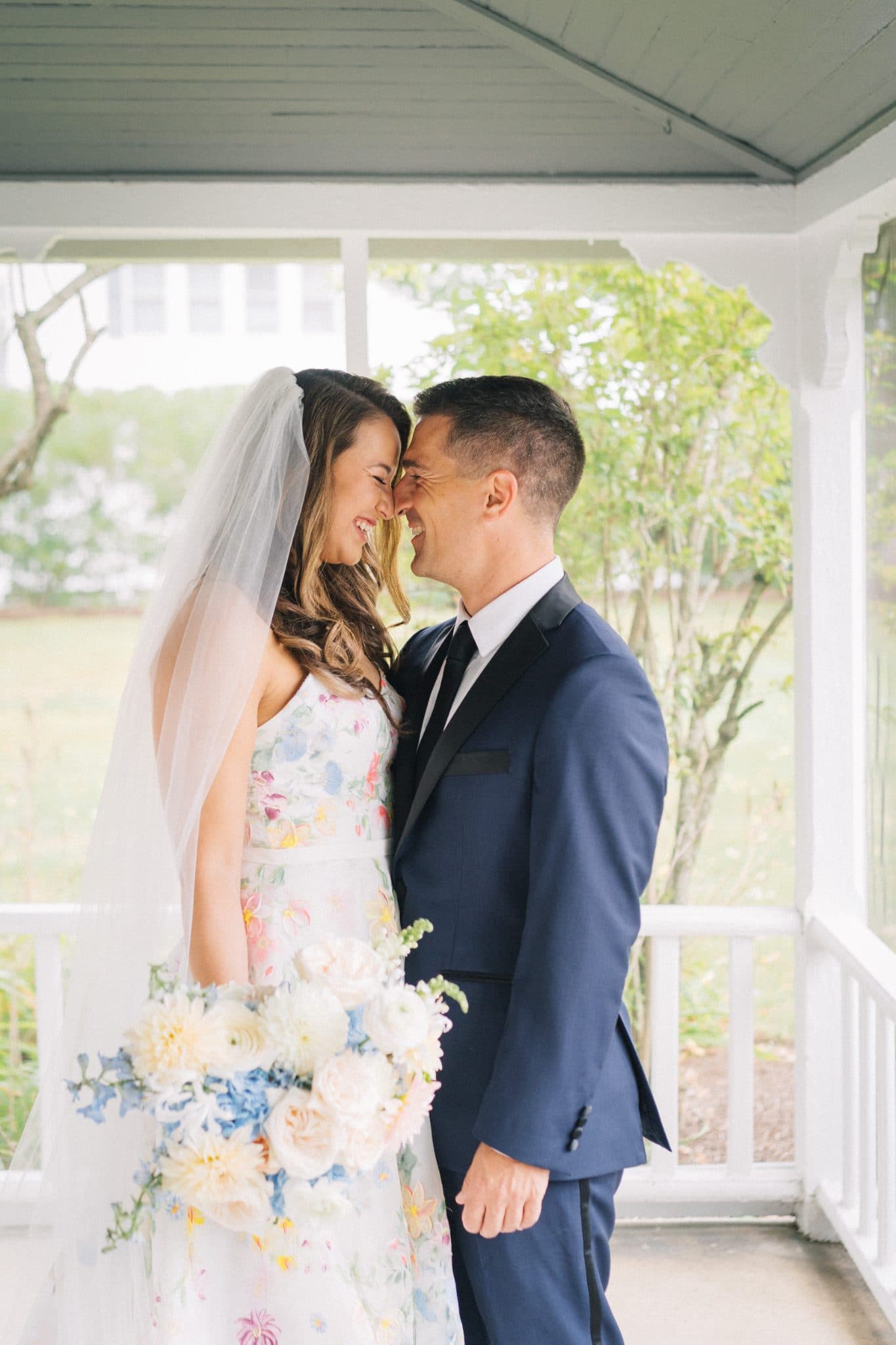 A bride in a floral dress and veil stands beside a man in a navy suit, both smiling and embracing on a porch.