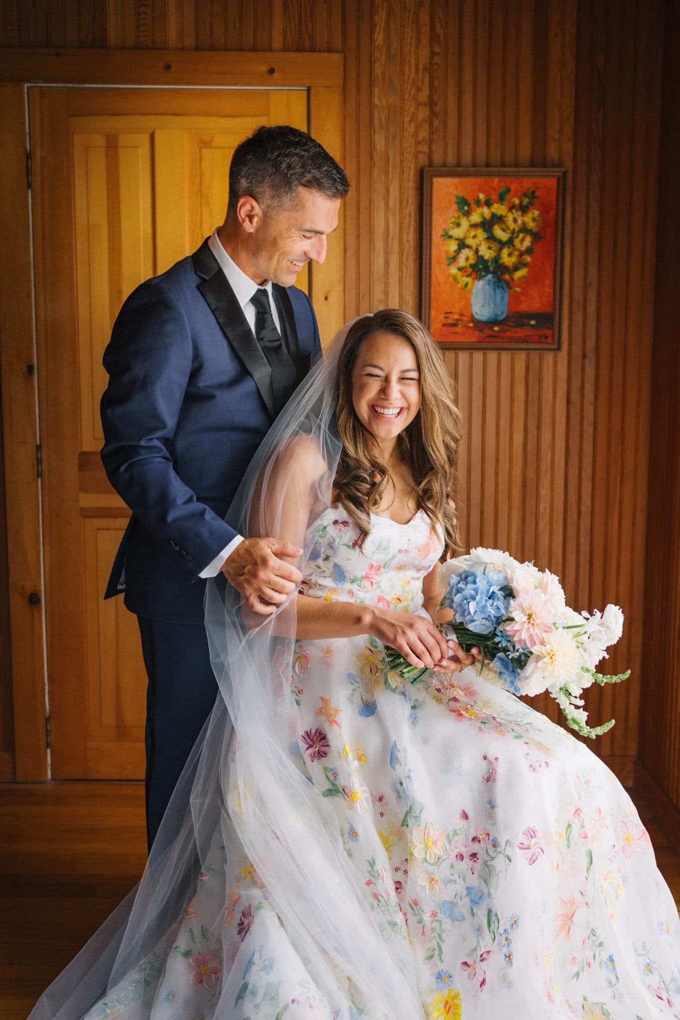 A bride in a floral wedding dress holds a bouquet, smiling, with a man in a suit standing beside her.