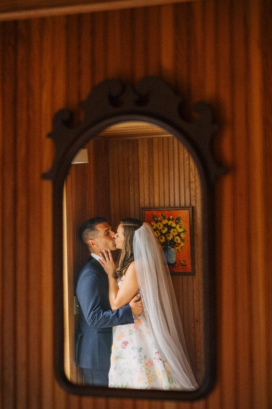 A couple kisses in a mirror reflecting a wooden-paneled wall with a floral painting and checkered curtains.