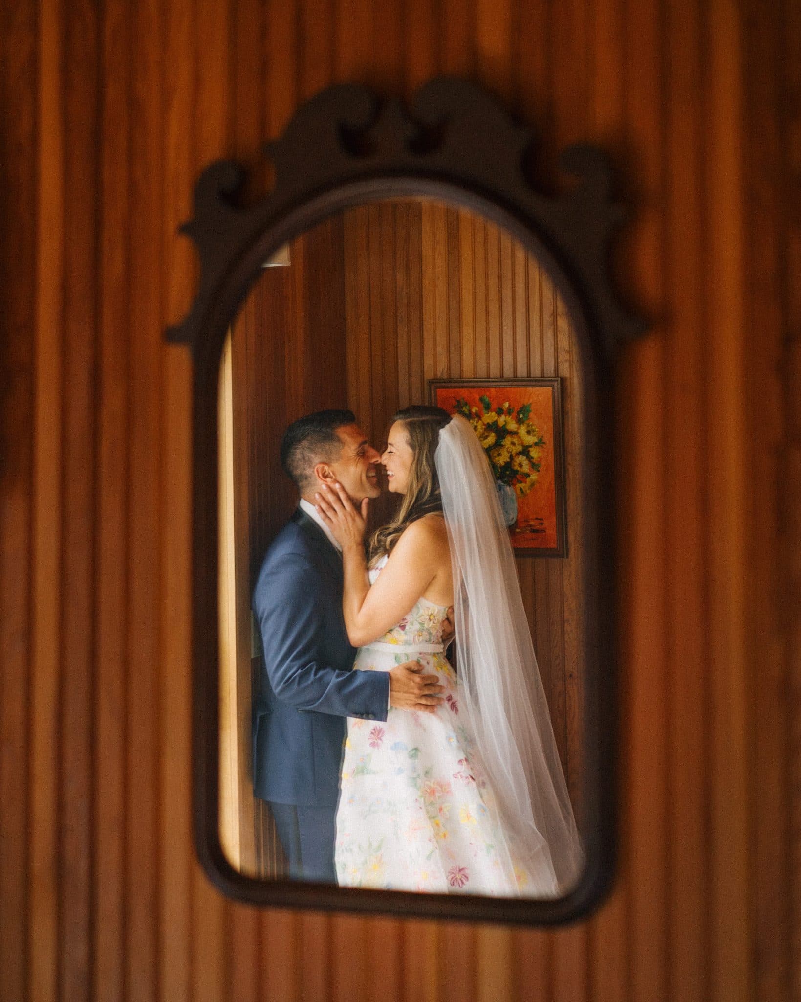 A couple in wedding attire is reflected in a mirror, embracing and smiling.