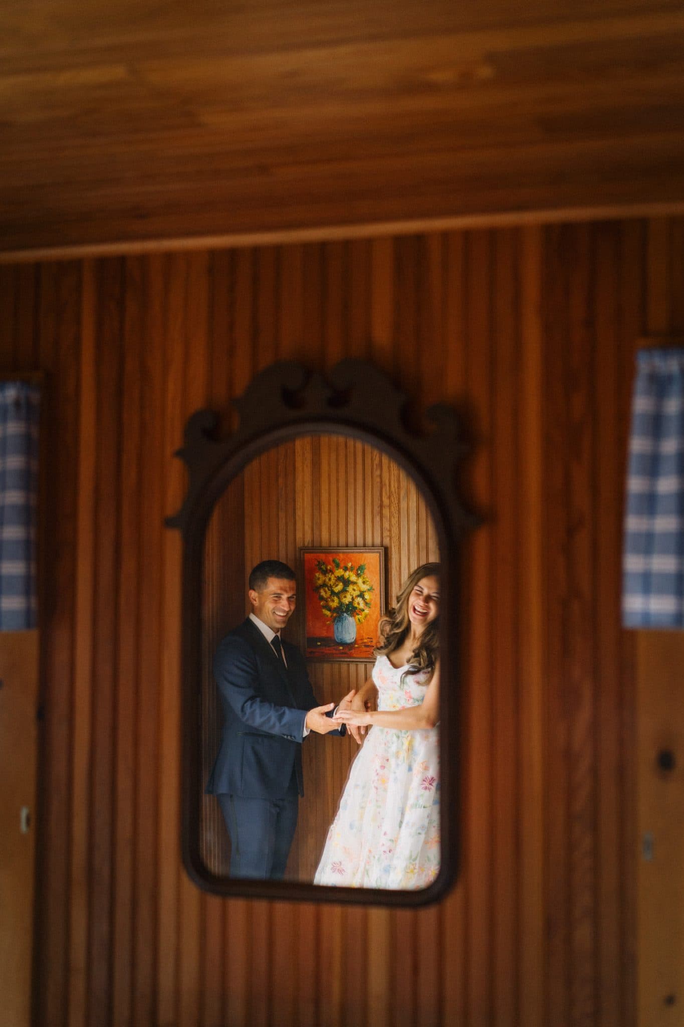A couple is reflected in a mirror, the man in a suit and the woman in a floral dress, both smiling.