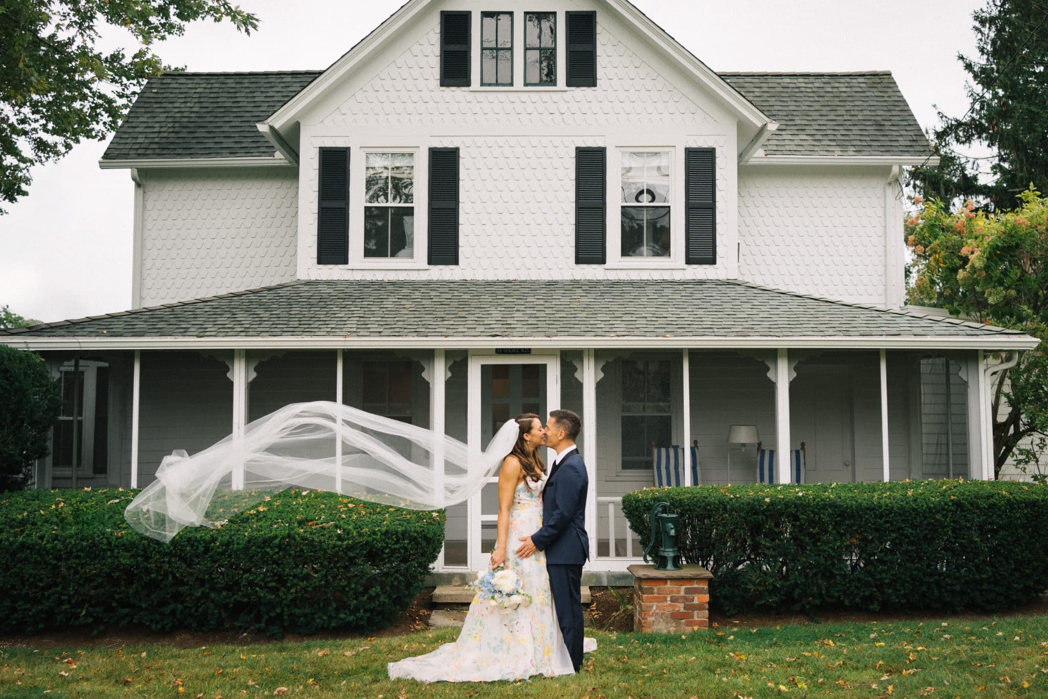 A bride and groom stand in front of a white house with a porch, the bride's veil flowing in the wind.