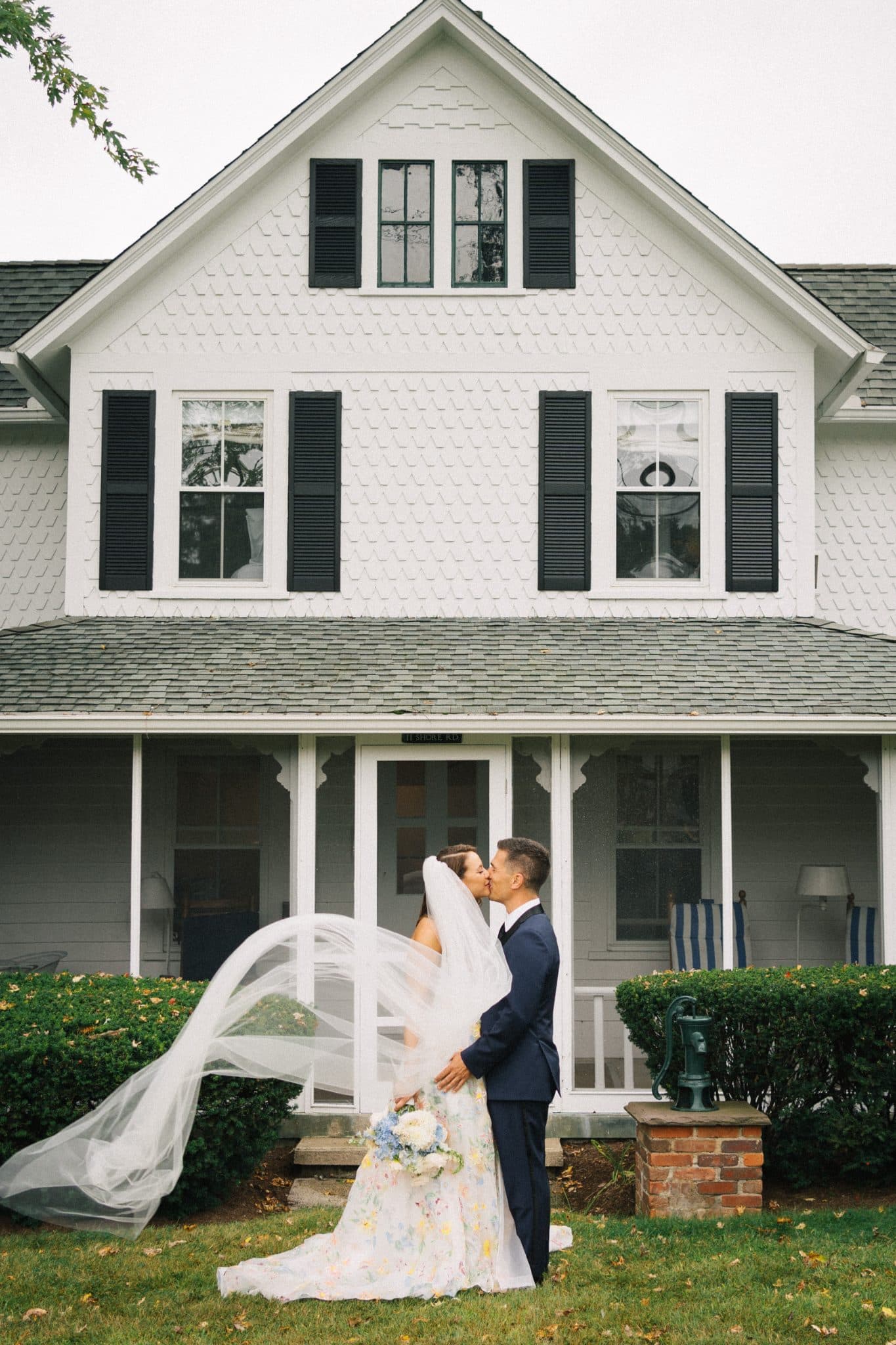 A bride and groom kiss in front of a white house with black shutters and a porch.