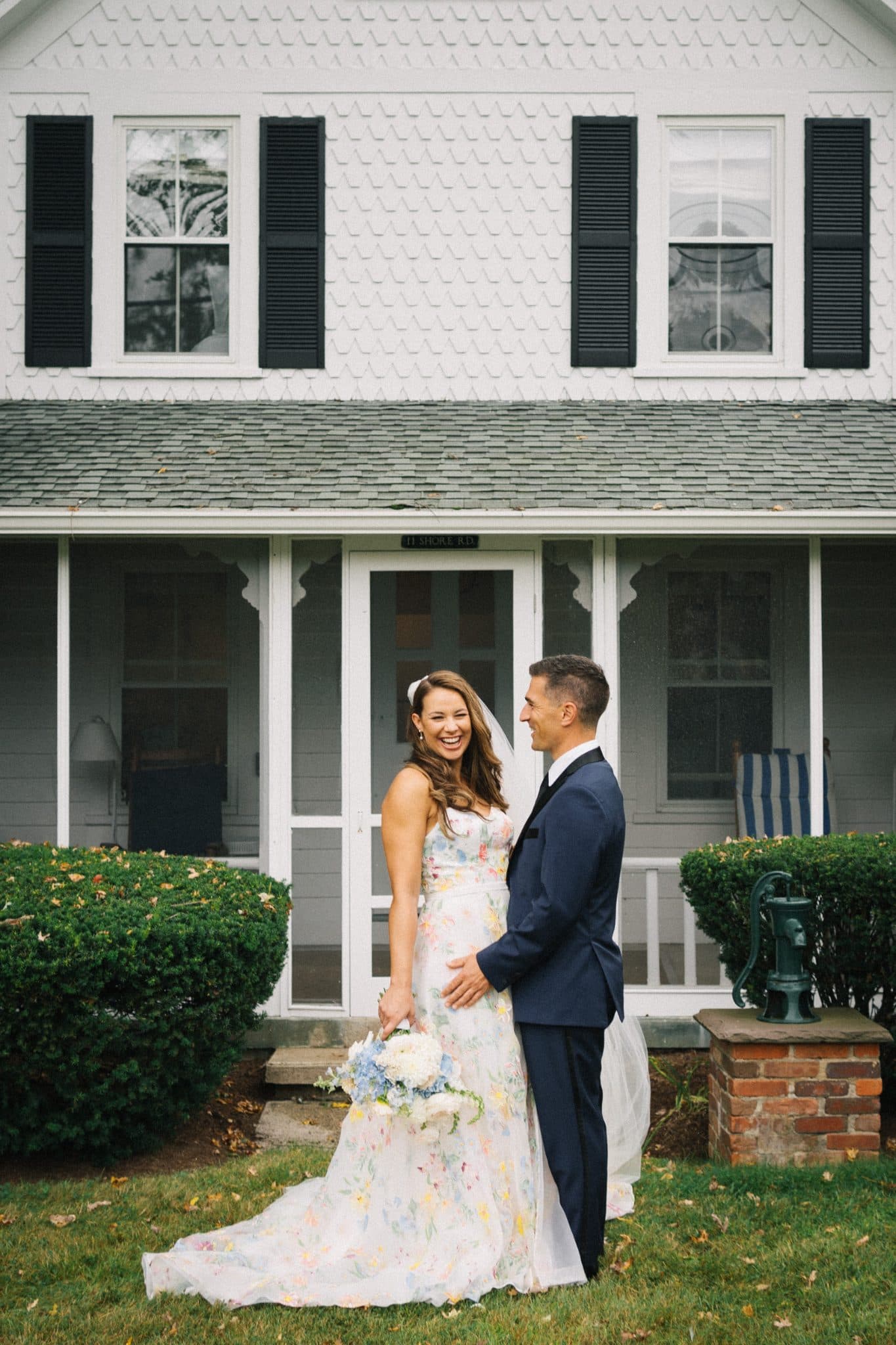 A bride in a floral dress and groom in a navy suit stand in front of a white house with black shutters and a porch.