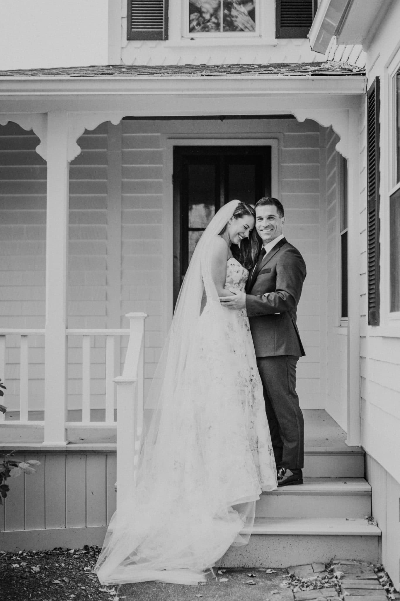 A bride and groom stand on a porch, embracing, with the bride wearing a long veil and the groom in a suit.