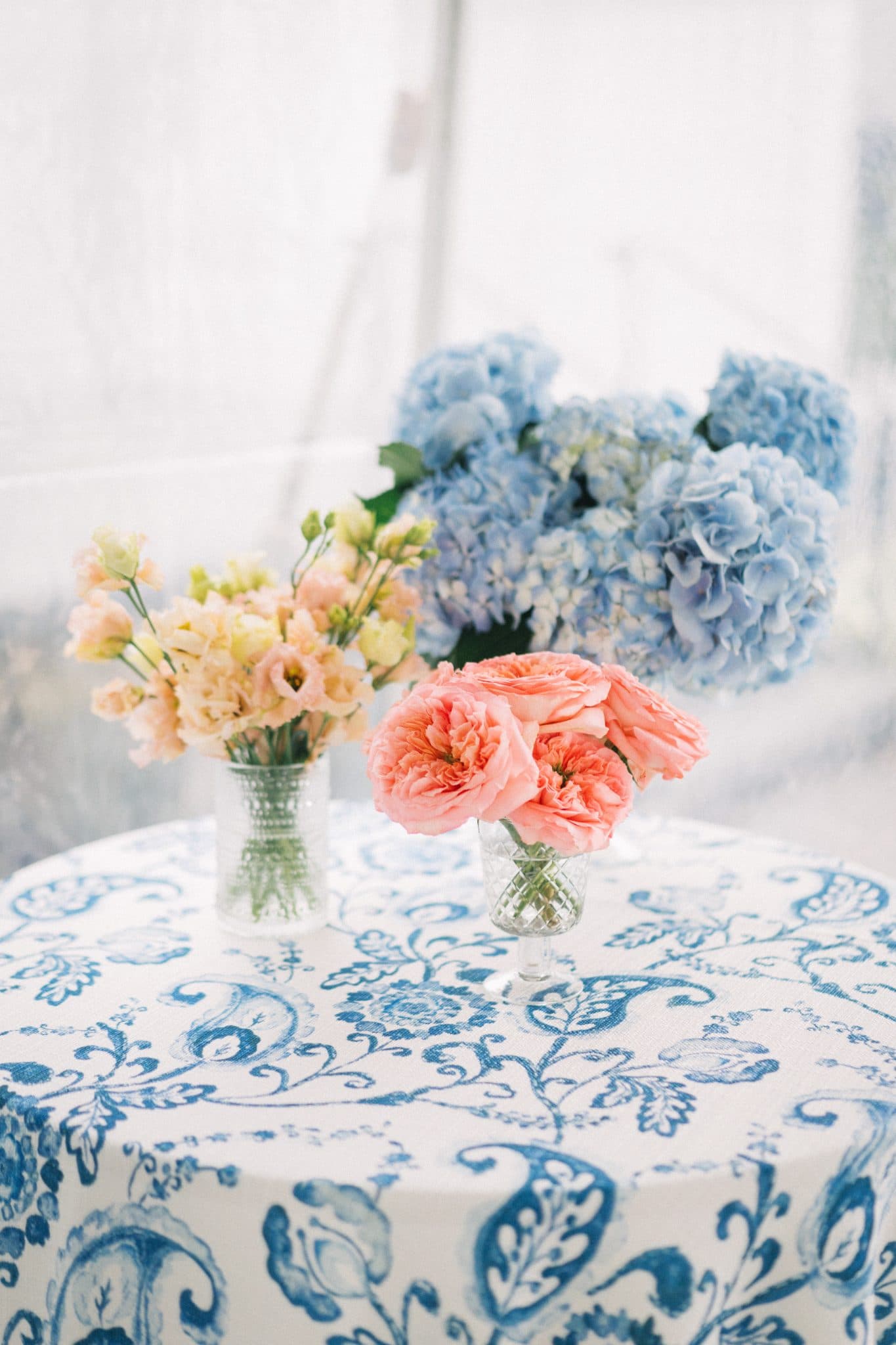 A table is adorned with a blue and white patterned tablecloth, featuring three vases of flowers: peach, yellow, and blue hydrangeas.
