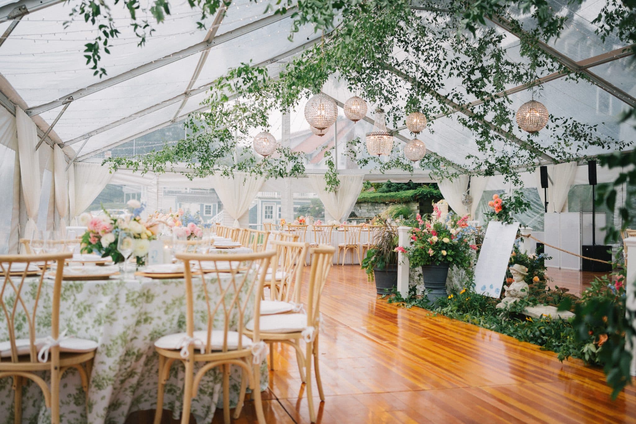 A beautifully decorated wedding tent with floral arrangements, hanging lights, and wooden chairs.