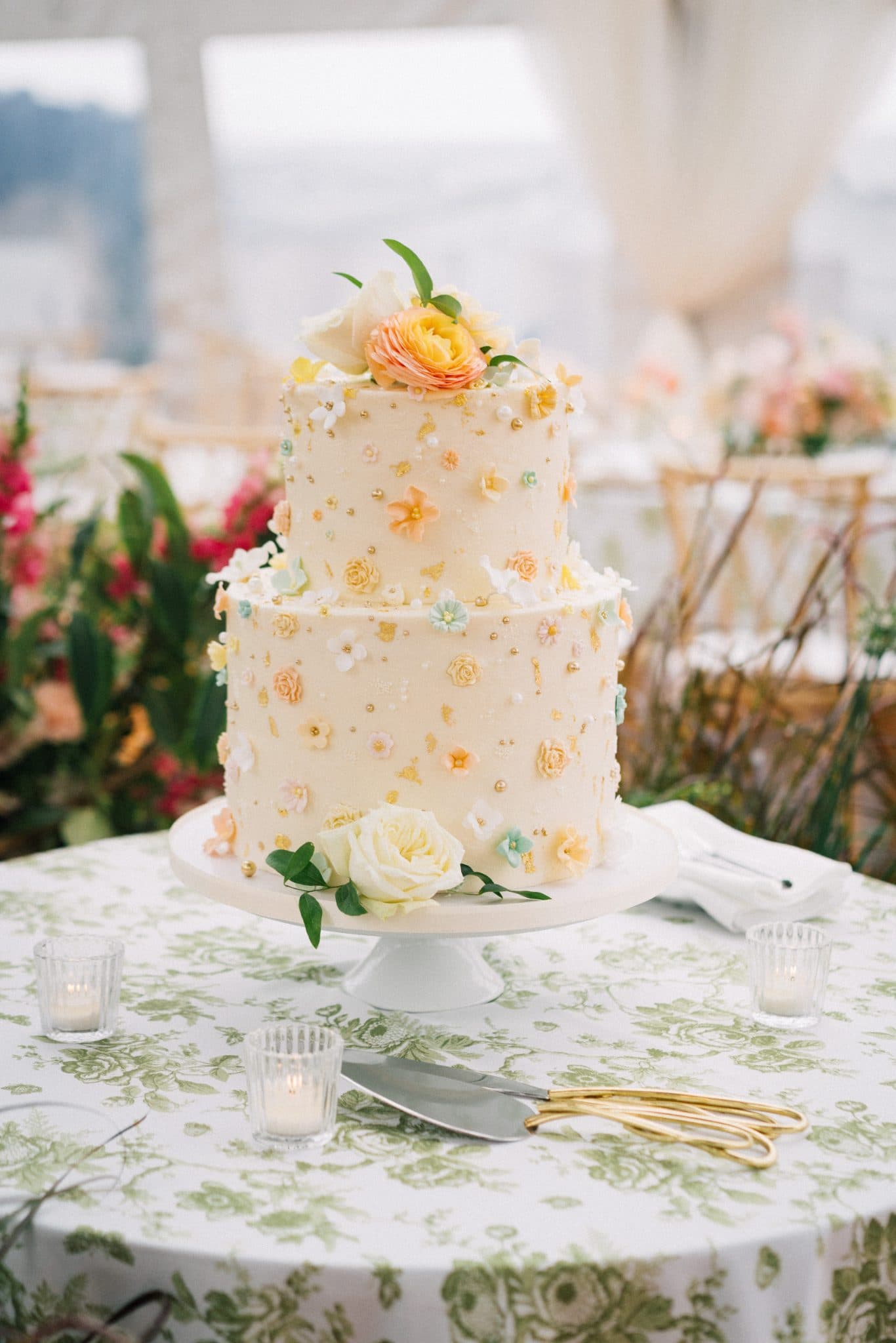 A two-tiered wedding cake adorned with flowers and small decorative elements sits on a floral-patterned tablecloth.