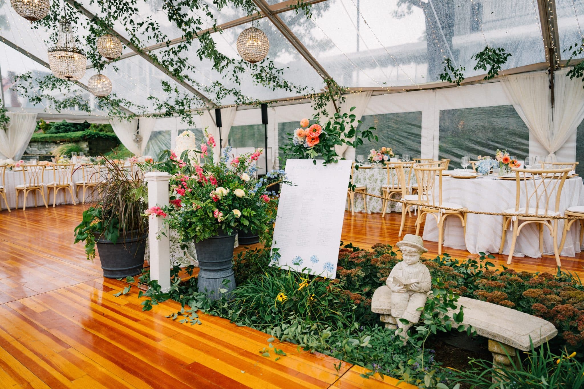A wedding reception inside a clear tent with floral arrangements, a statue, and a cake in the background.
