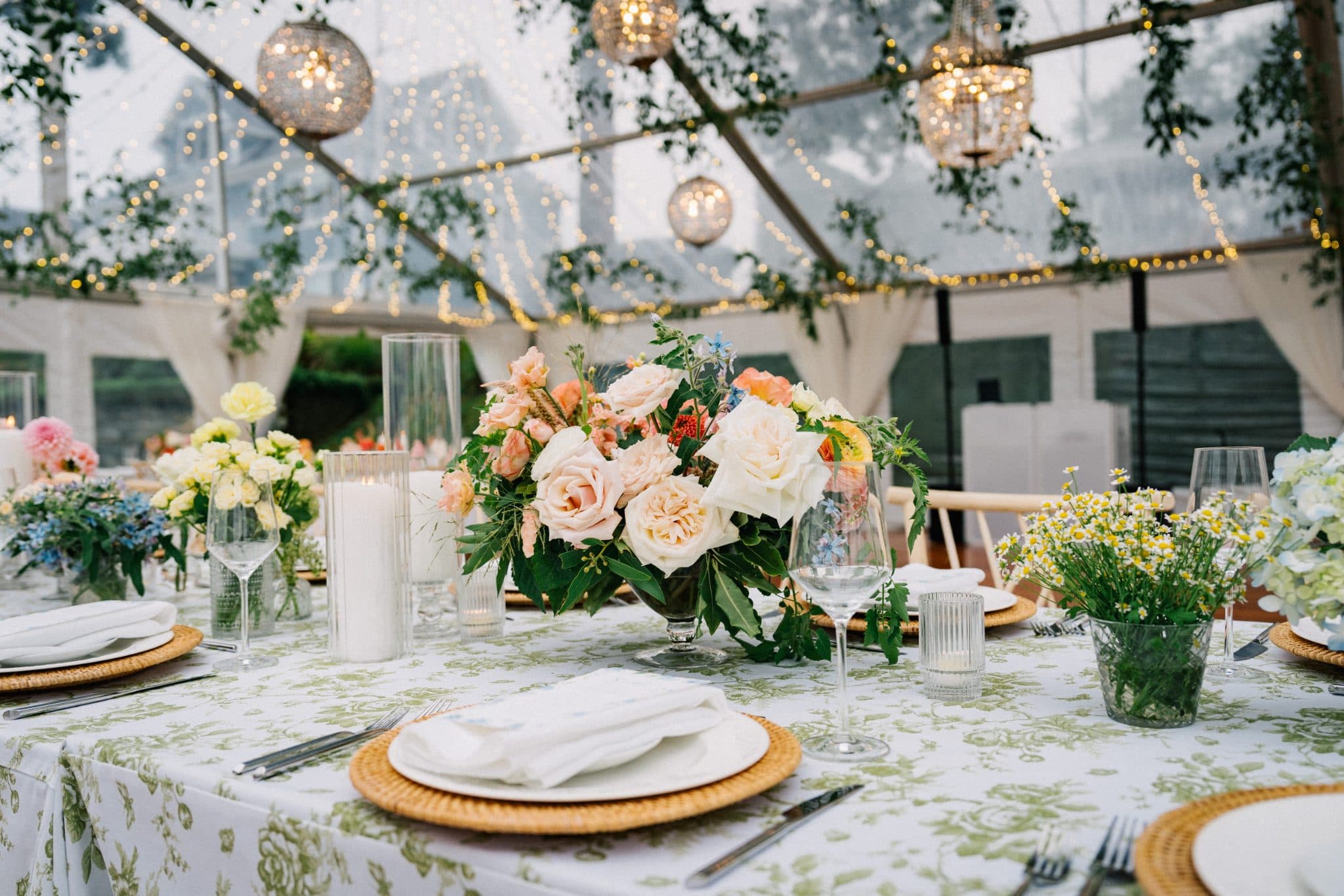 A beautifully set table with floral centerpieces, candles, and elegant tableware under a tent adorned with string lights and greenery.