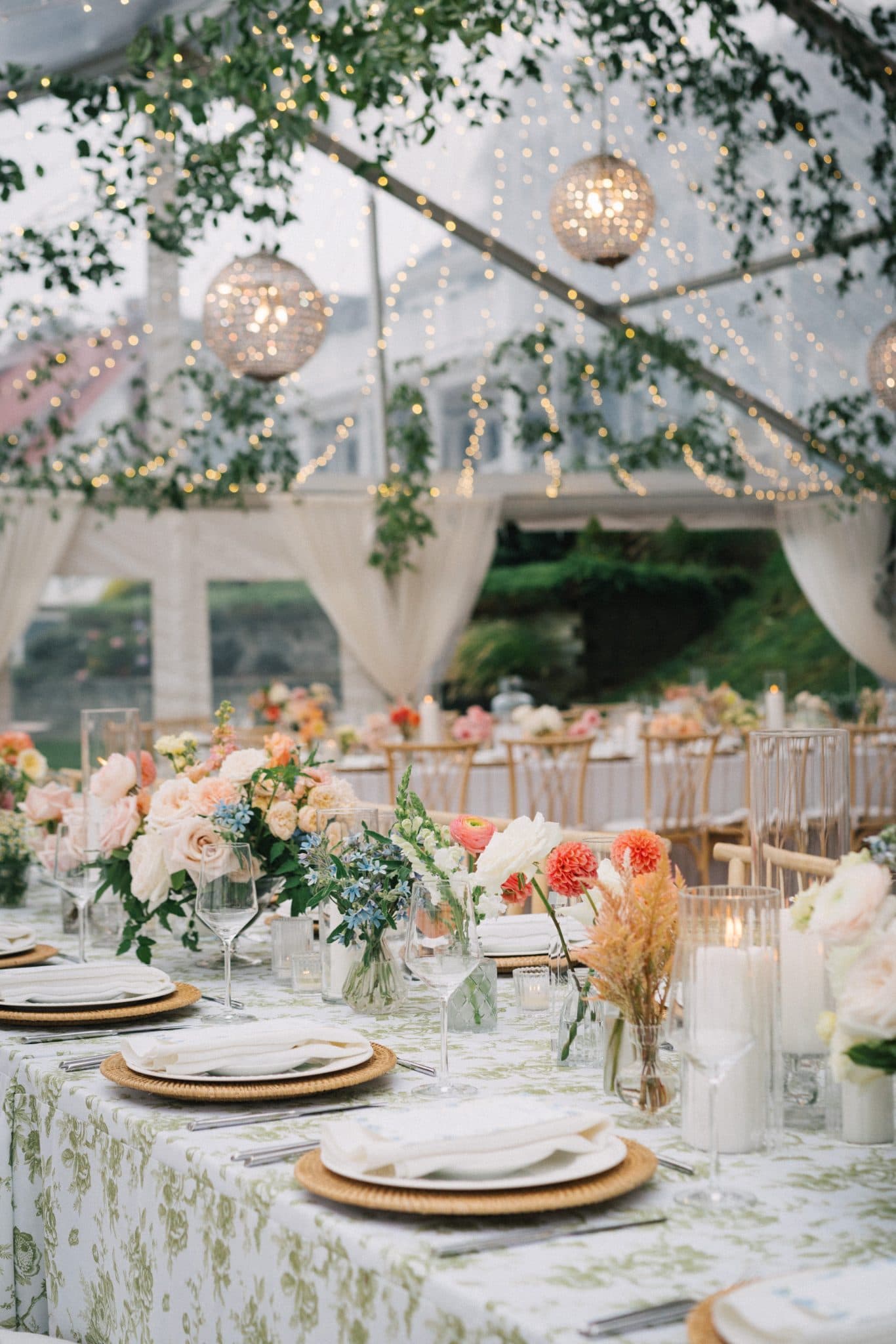 A beautifully set wedding table adorned with floral arrangements, candles, and elegant tableware under a tent decorated with string lights and greenery.