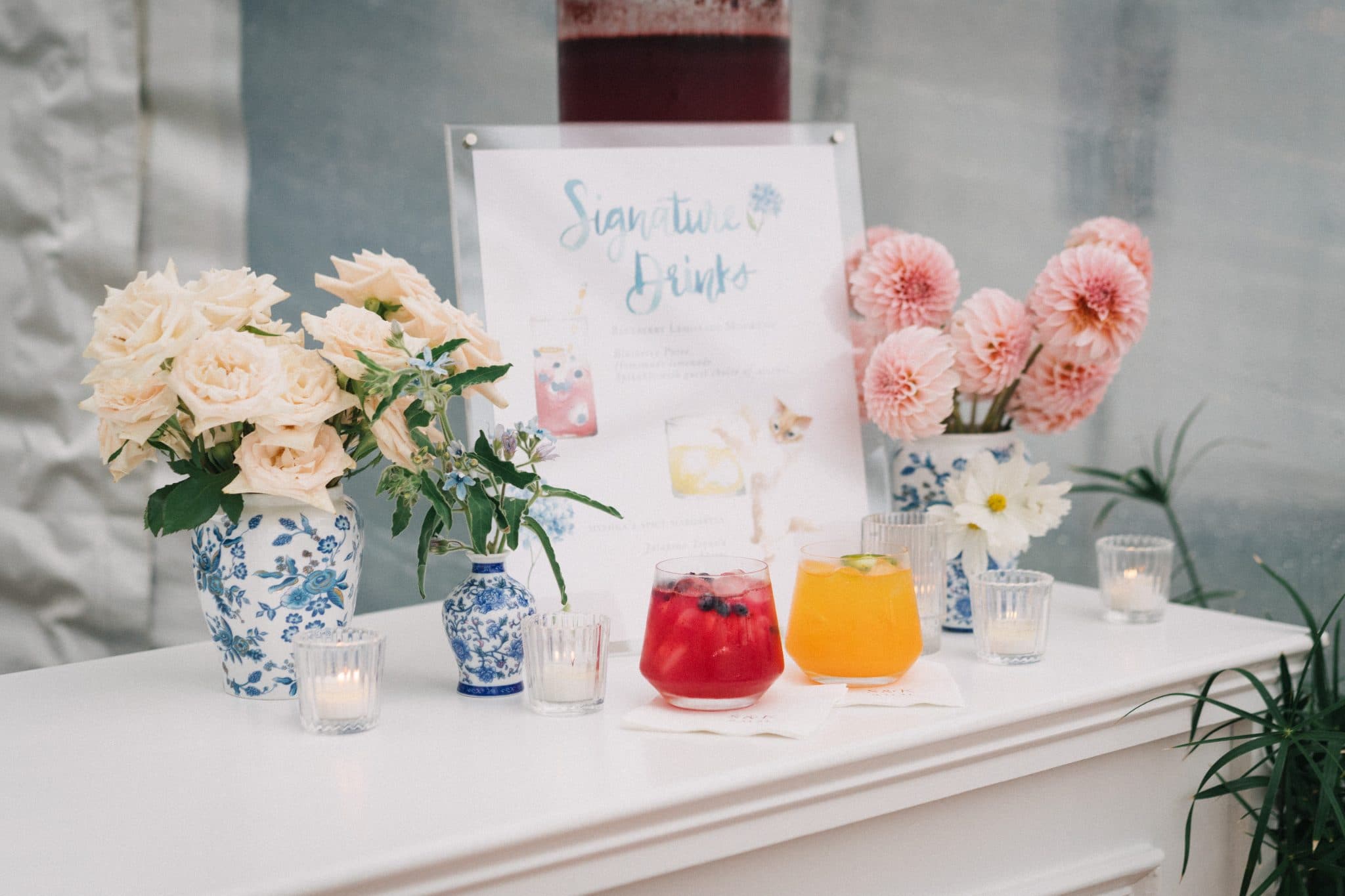 A wedding bar setup features two colorful drinks, vases with white and pink flowers, candles, and a sign for signature drinks.