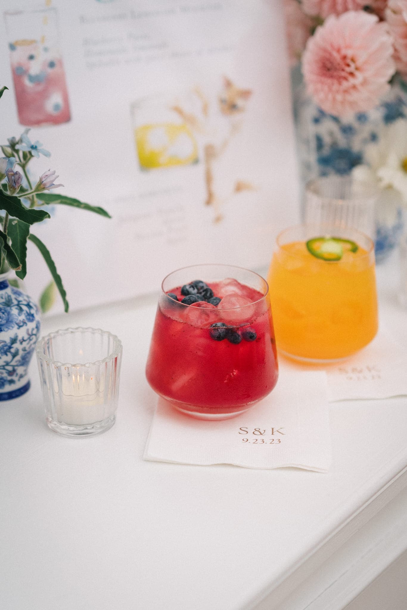 A wedding table setting features a red drink with blueberries, a yellow drink with a jalapeño slice, a lit candle, and a napkin with initials and a date.