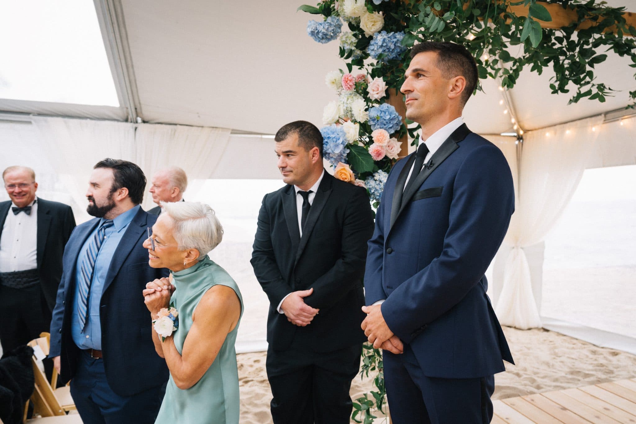 A wedding scene with a bride in a white dress, groom in a dark suit, and guests in formal attire under a floral arch.