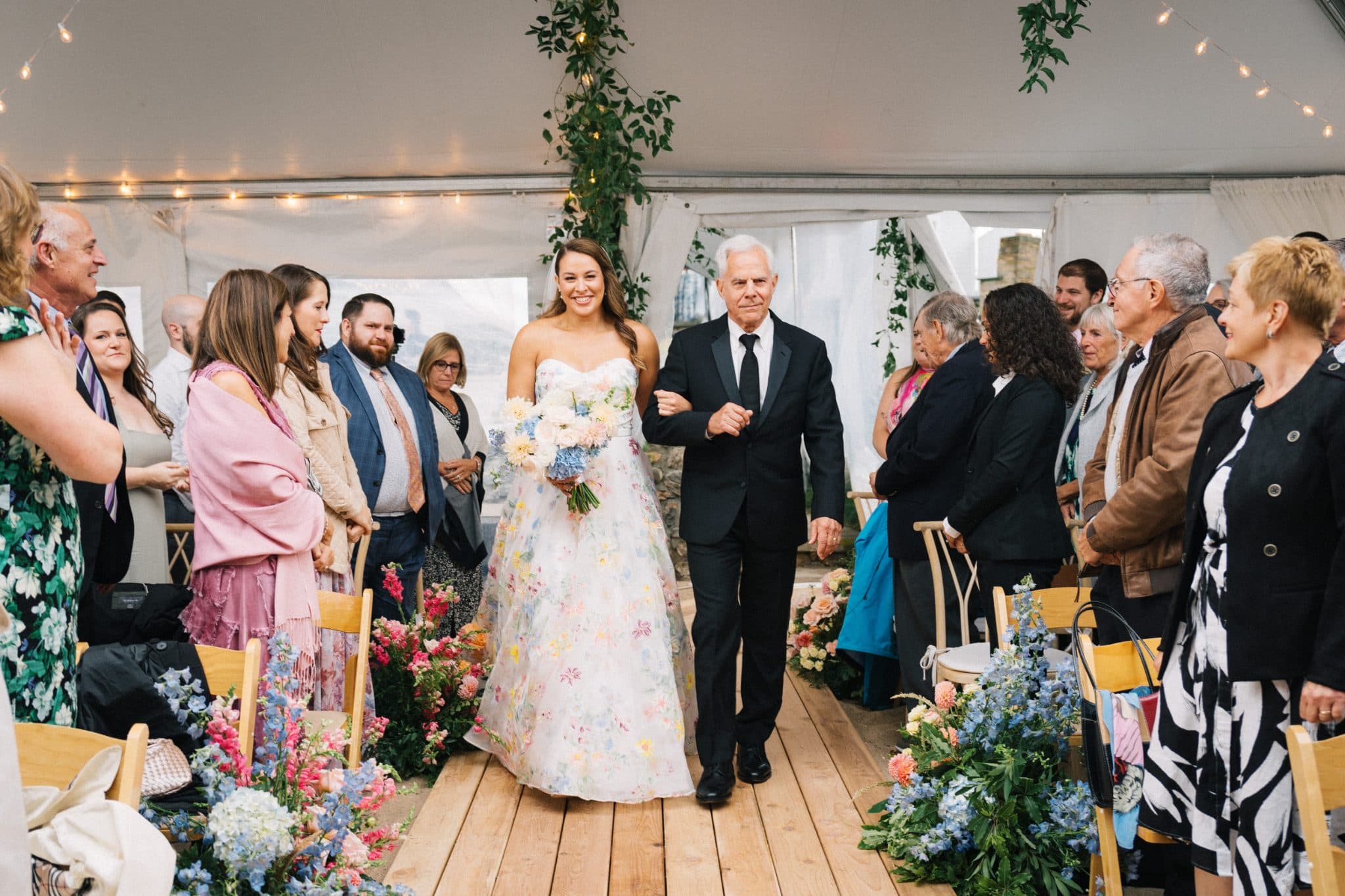 A bride in a floral dress walks down an aisle with her father, surrounded by guests in a tent adorned with flowers and string lights.