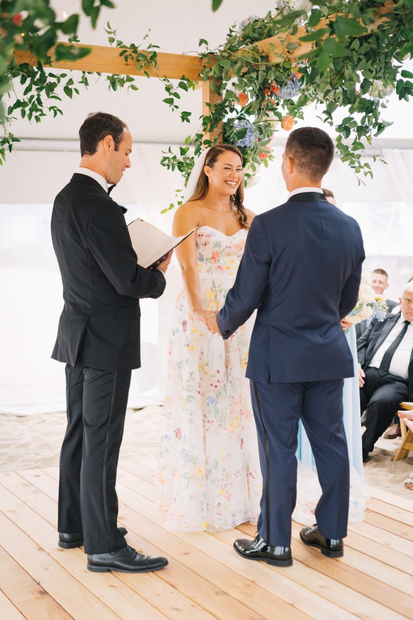 A bride in a floral dress holds hands with a groom in a navy suit under a floral archway during a wedding ceremony.