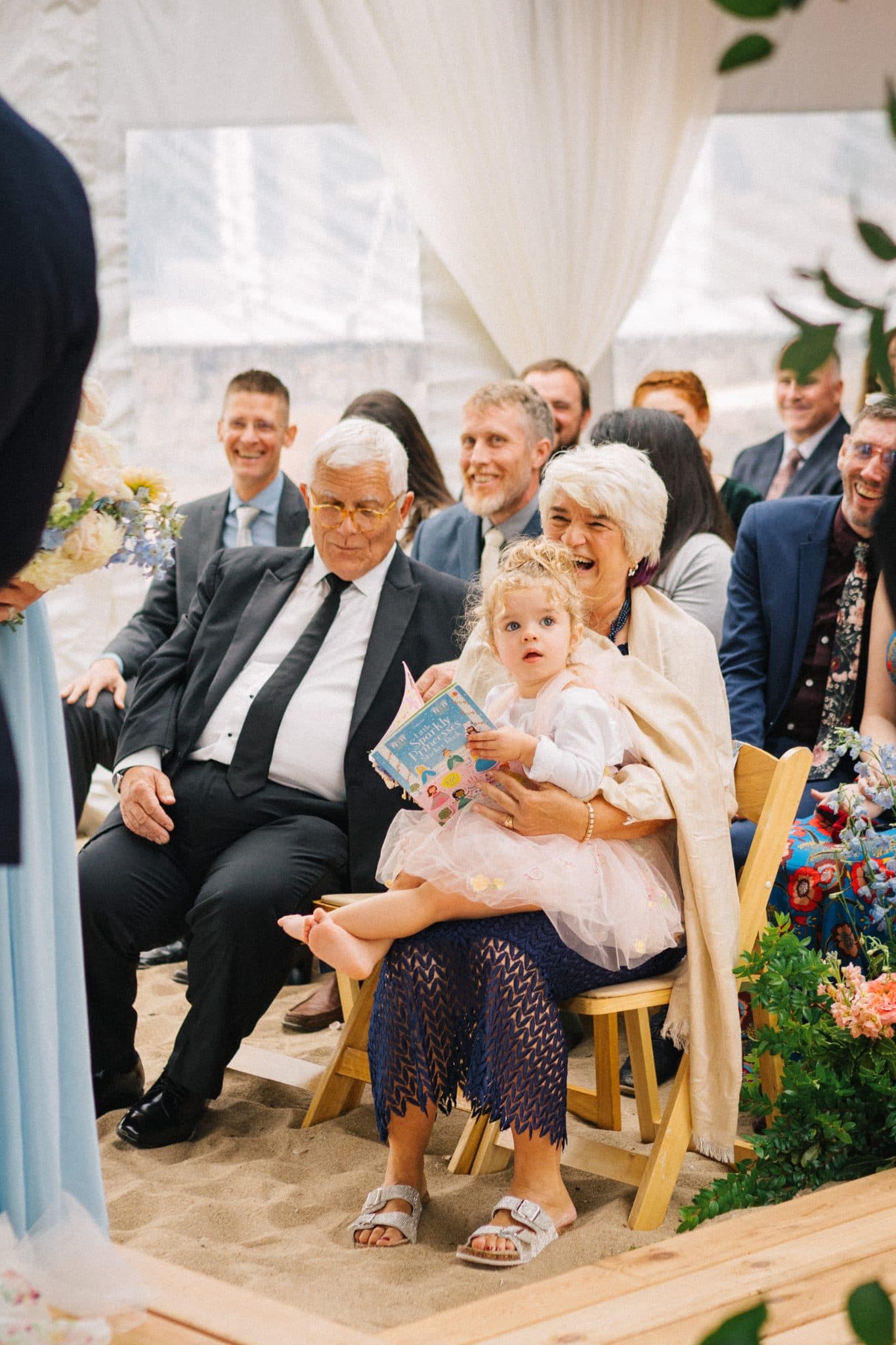 A woman holds a young girl in a wedding ceremony, surrounded by seated guests and floral decorations.