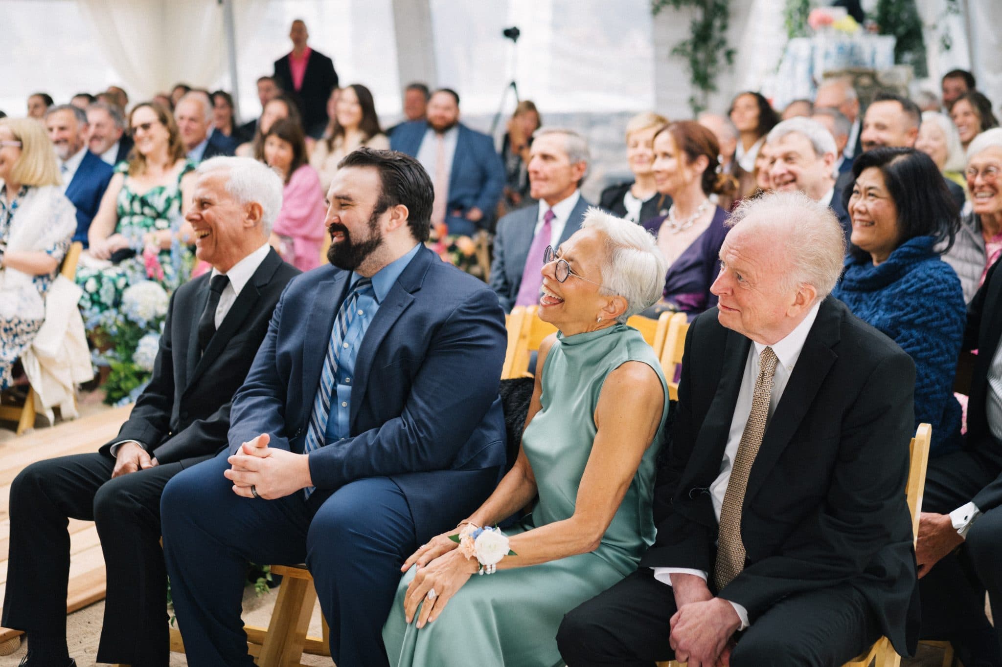 A group of people in formal attire are seated on wooden chairs, smiling and laughing during a wedding ceremony.