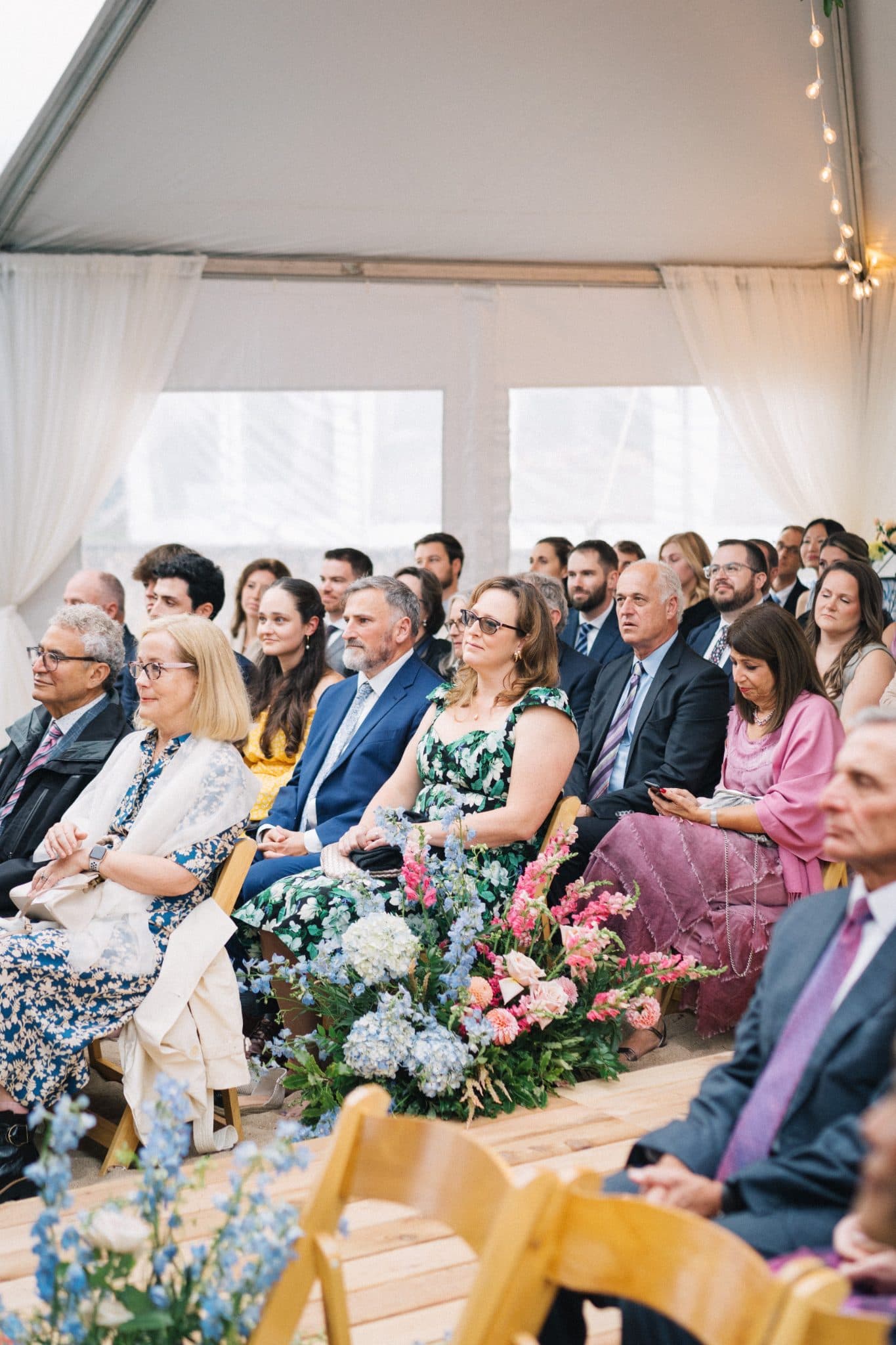 A group of people seated in chairs under a tent, surrounded by floral arrangements, attentively watching a ceremony.