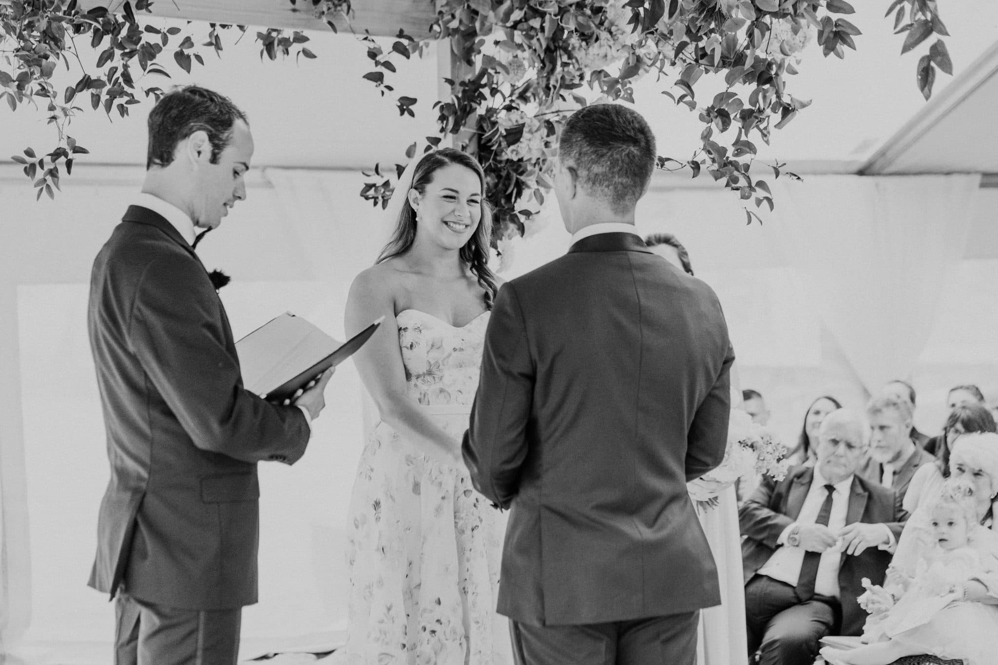 A bride in a floral dress stands between a groom in a suit and a officiant holding a book, with guests seated in the background.