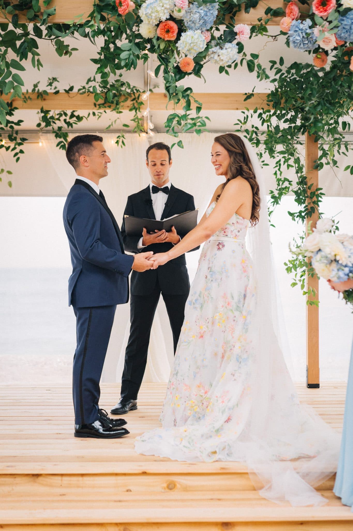 A bride in a floral dress and a groom in a blue suit stand at an altar under a floral arch, exchanging vows with a officiant.