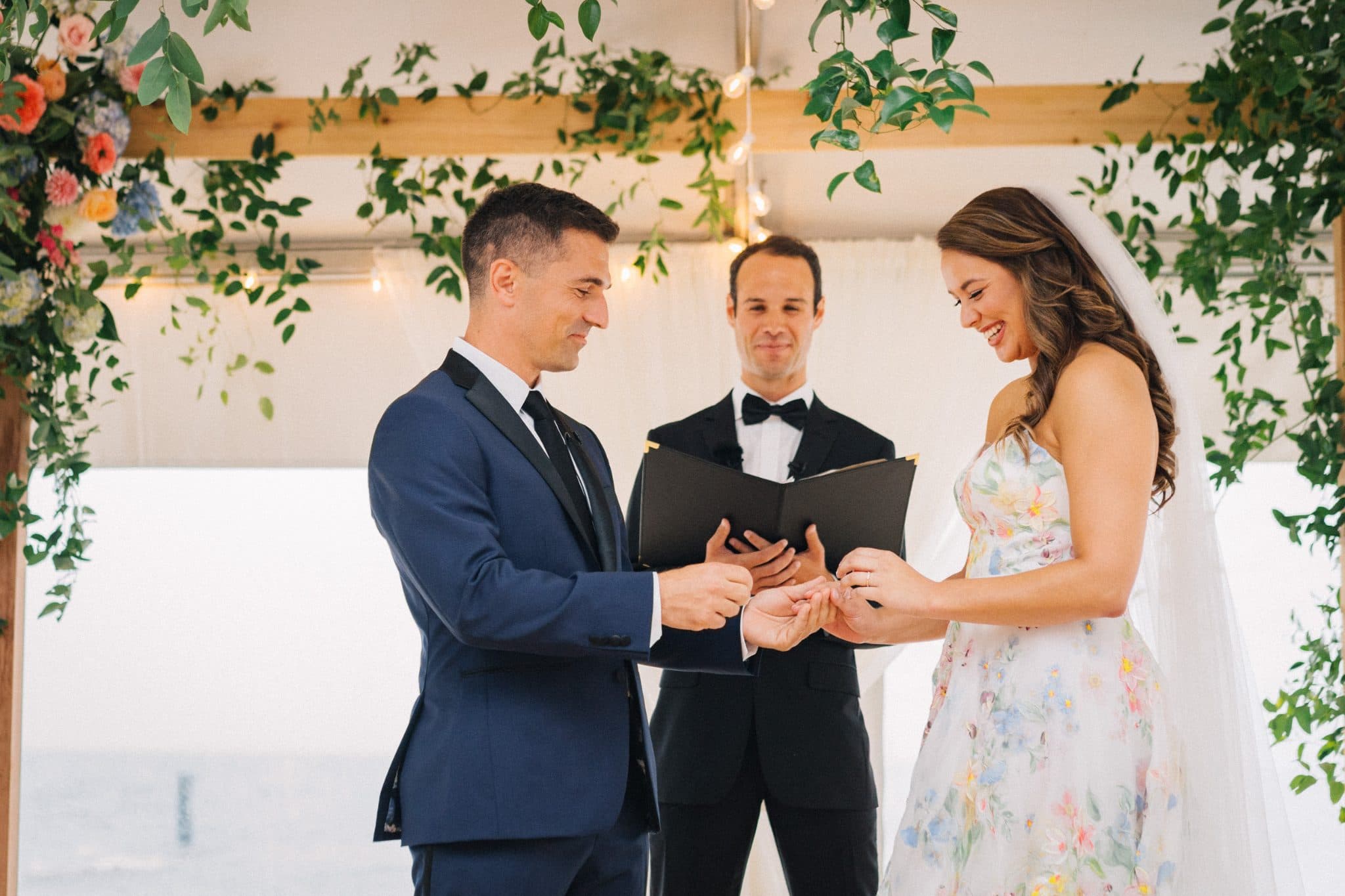 A bride in a floral dress and a groom in a blue suit exchange rings during a wedding ceremony under a floral archway.