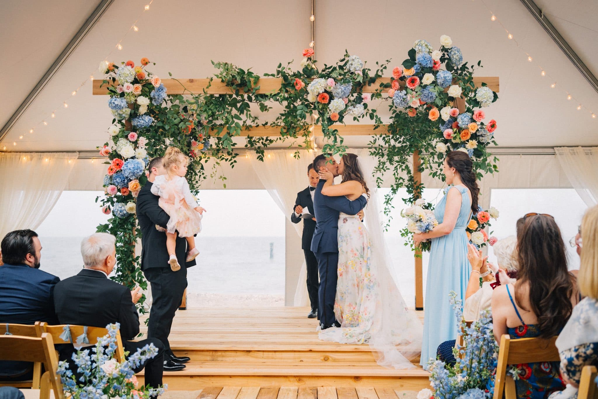 A couple kisses under a floral archway at a wedding ceremony, with guests seated and a ring bearer holding a child.