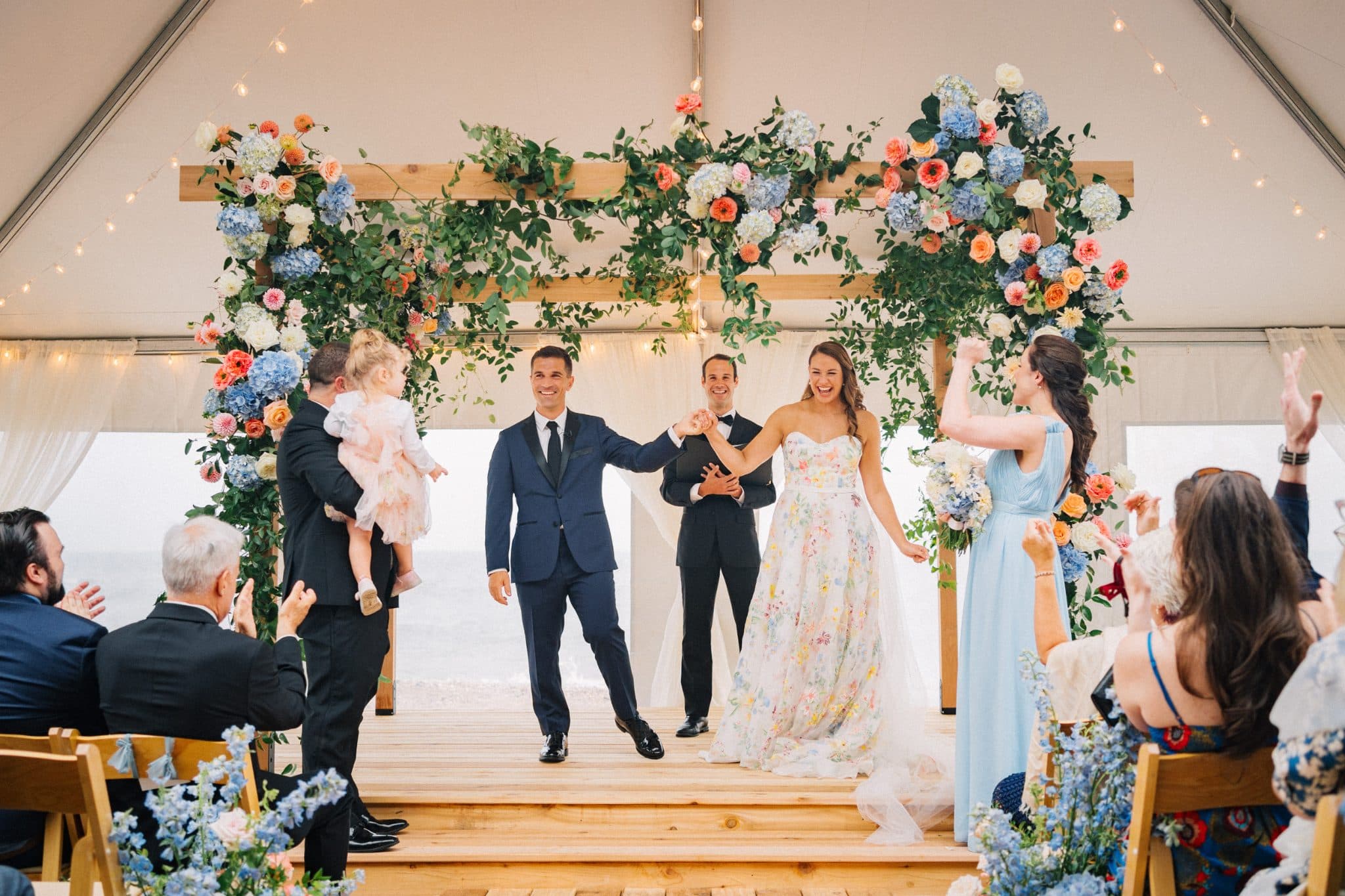 A bride in a floral dress and groom in a suit walk down a wooden aisle under a floral archway, surrounded by guests clapping.