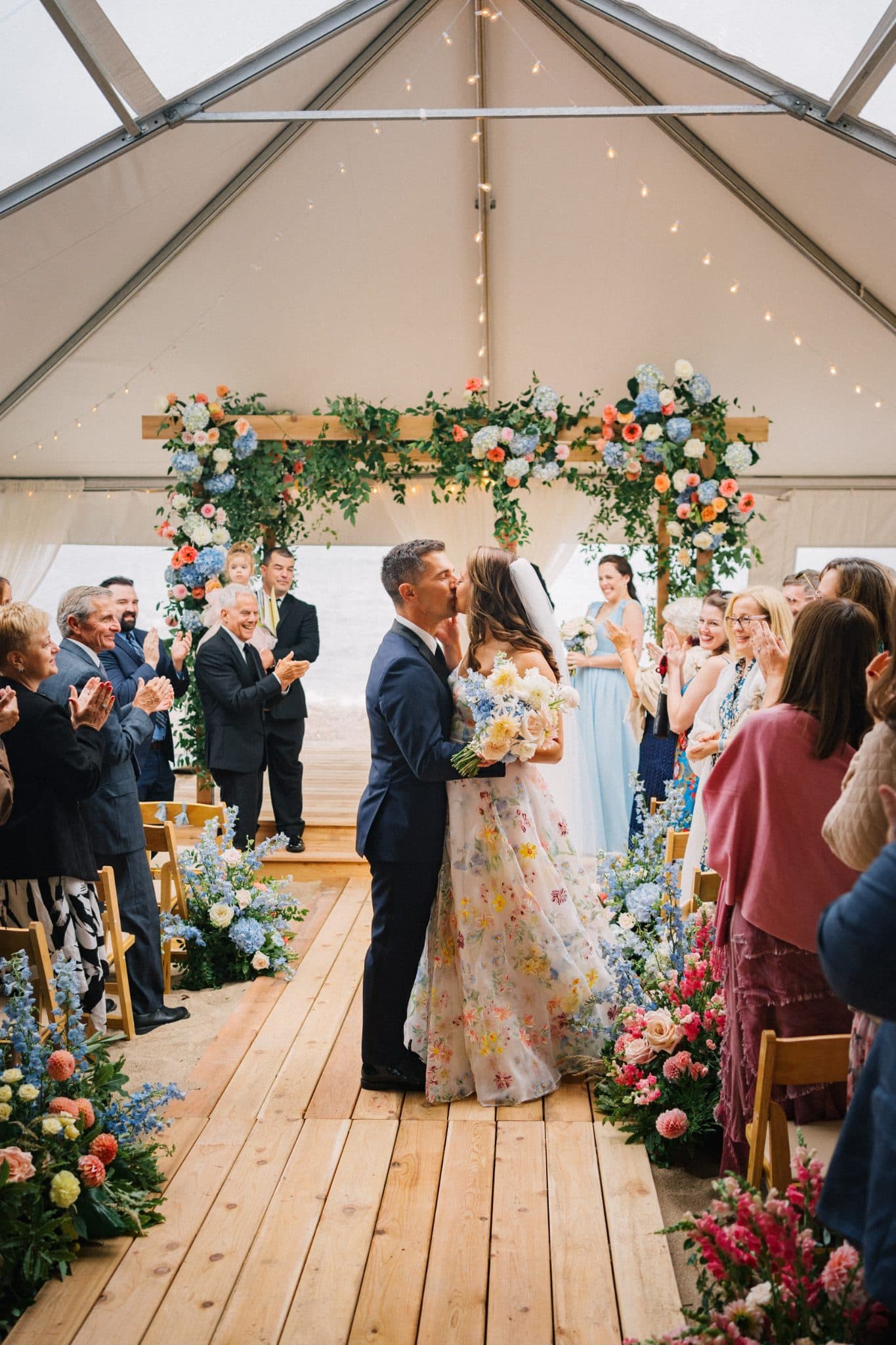 A couple kisses under a floral archway at a wedding ceremony, surrounded by guests clapping.