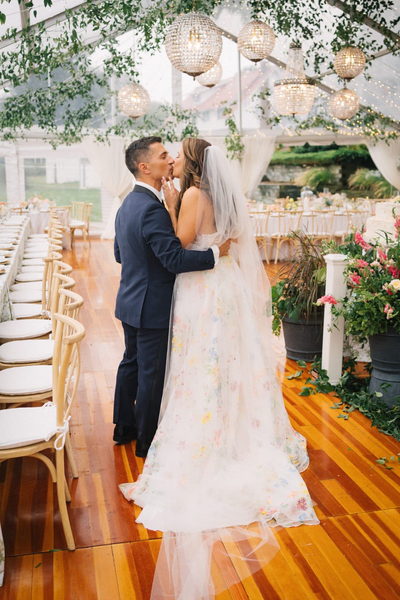 A bride in a floral dress and veil kisses a groom in a dark suit under a tent with hanging lights and greenery.