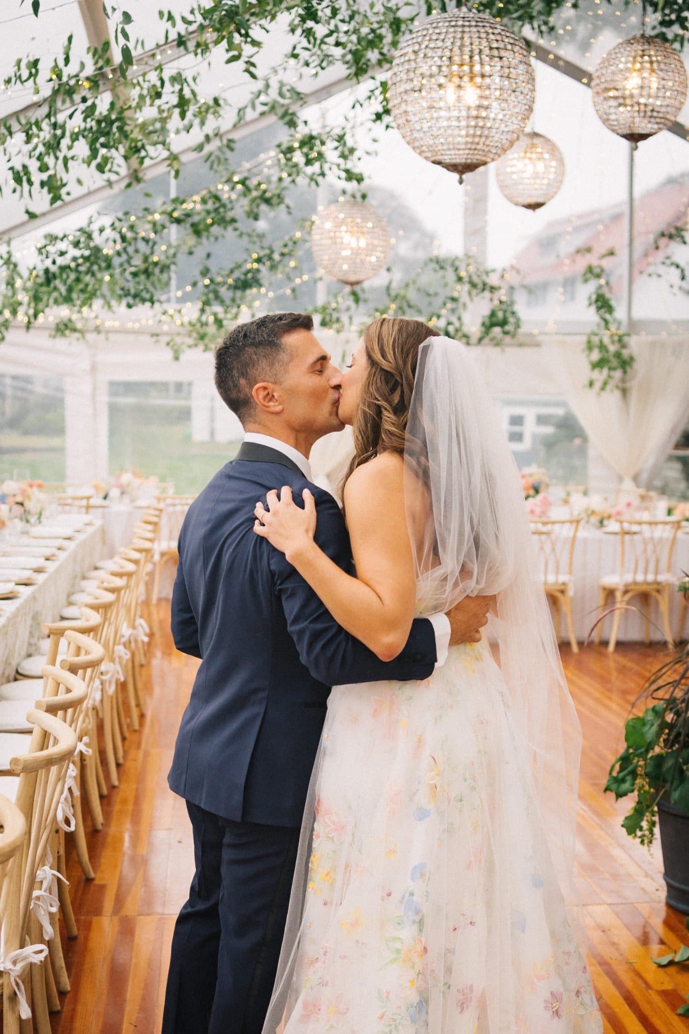 A bride in a floral gown and veil kisses a groom in a navy suit under a canopy of greenery and sparkling lights.