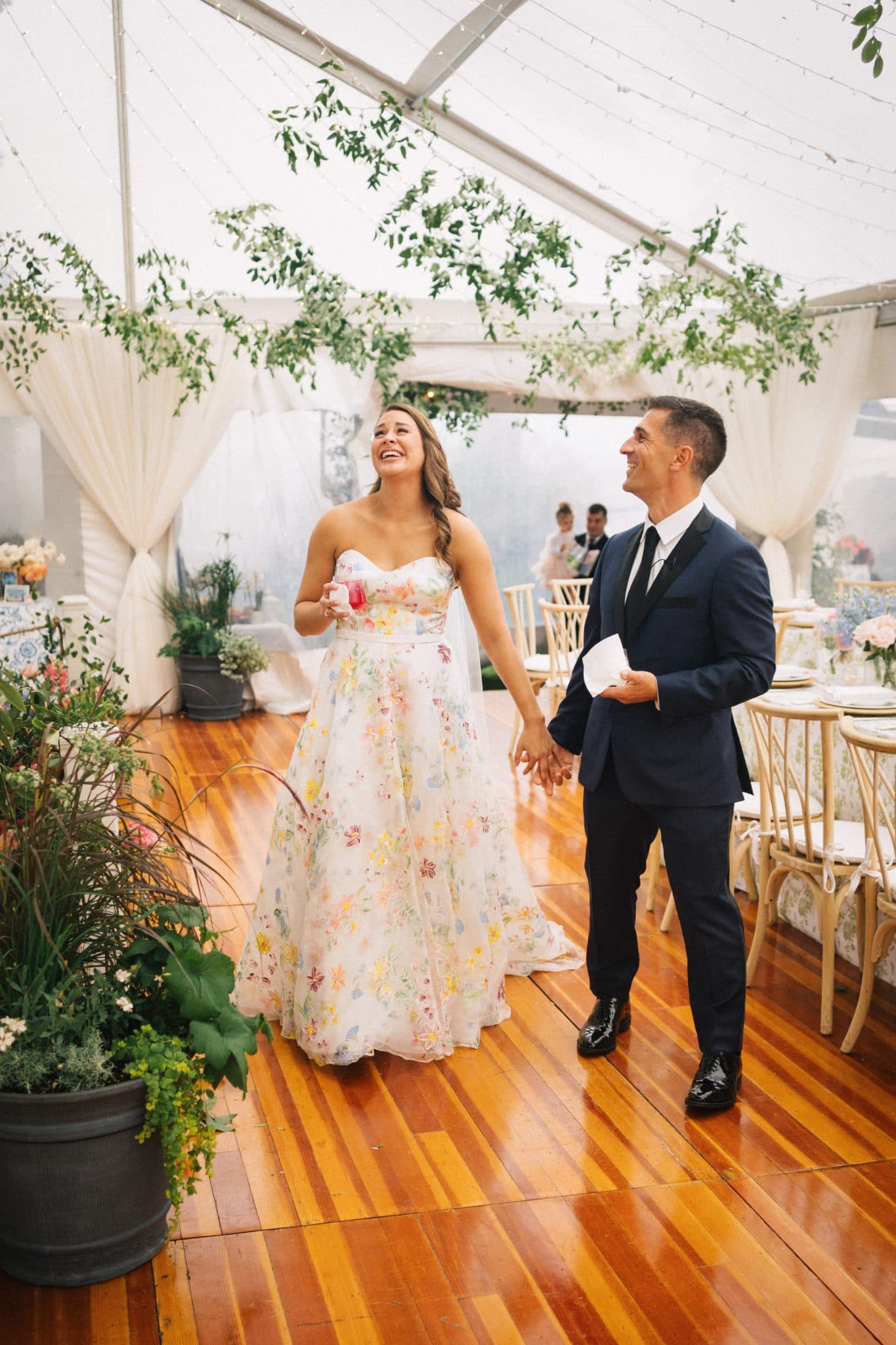 A couple walks hand in hand under a white canopy, surrounded by greenery and wooden furniture.