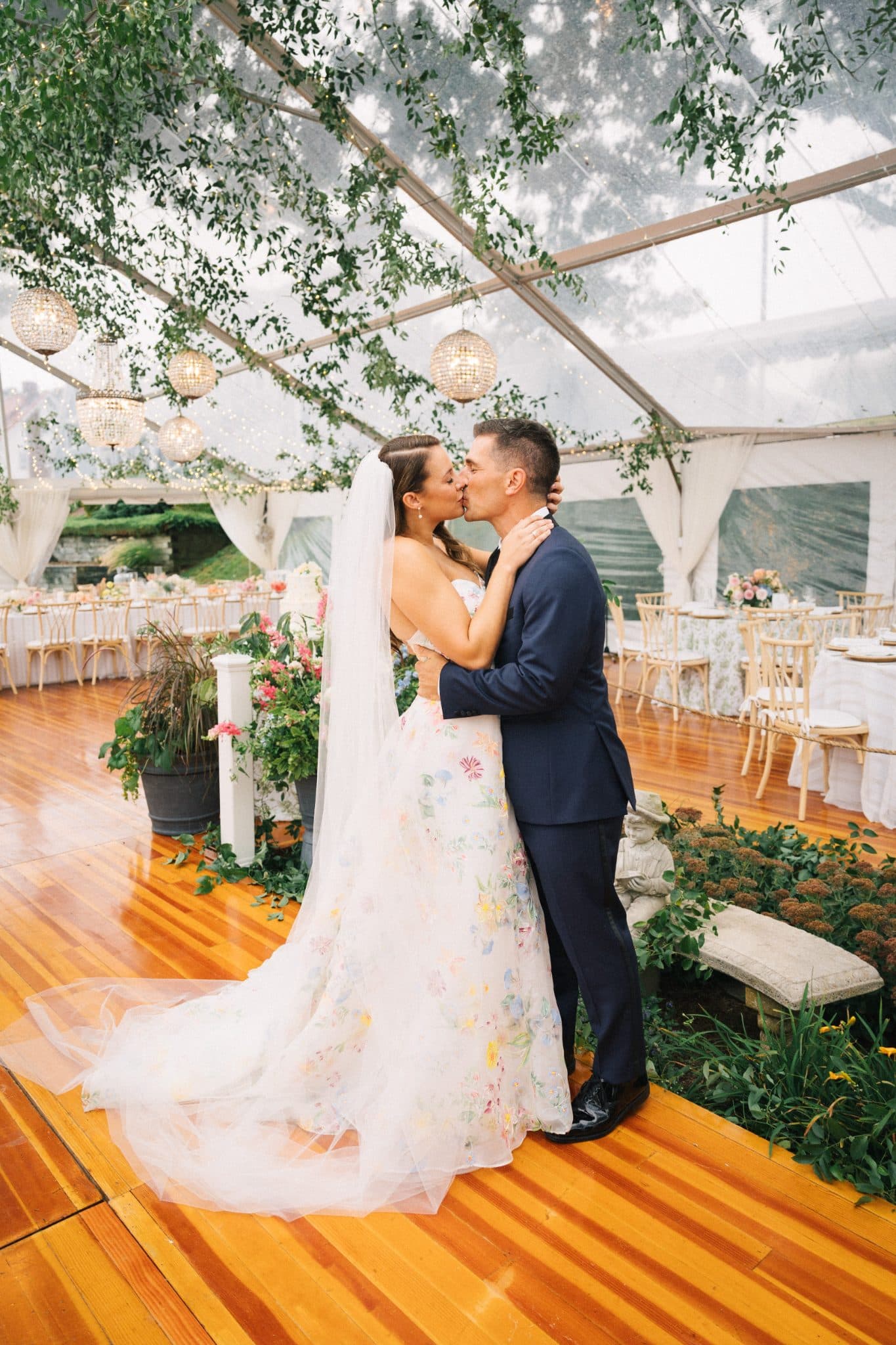 A bride in a floral dress and veil kisses a groom in a dark suit under a clear tent adorned with greenery and hanging lights.