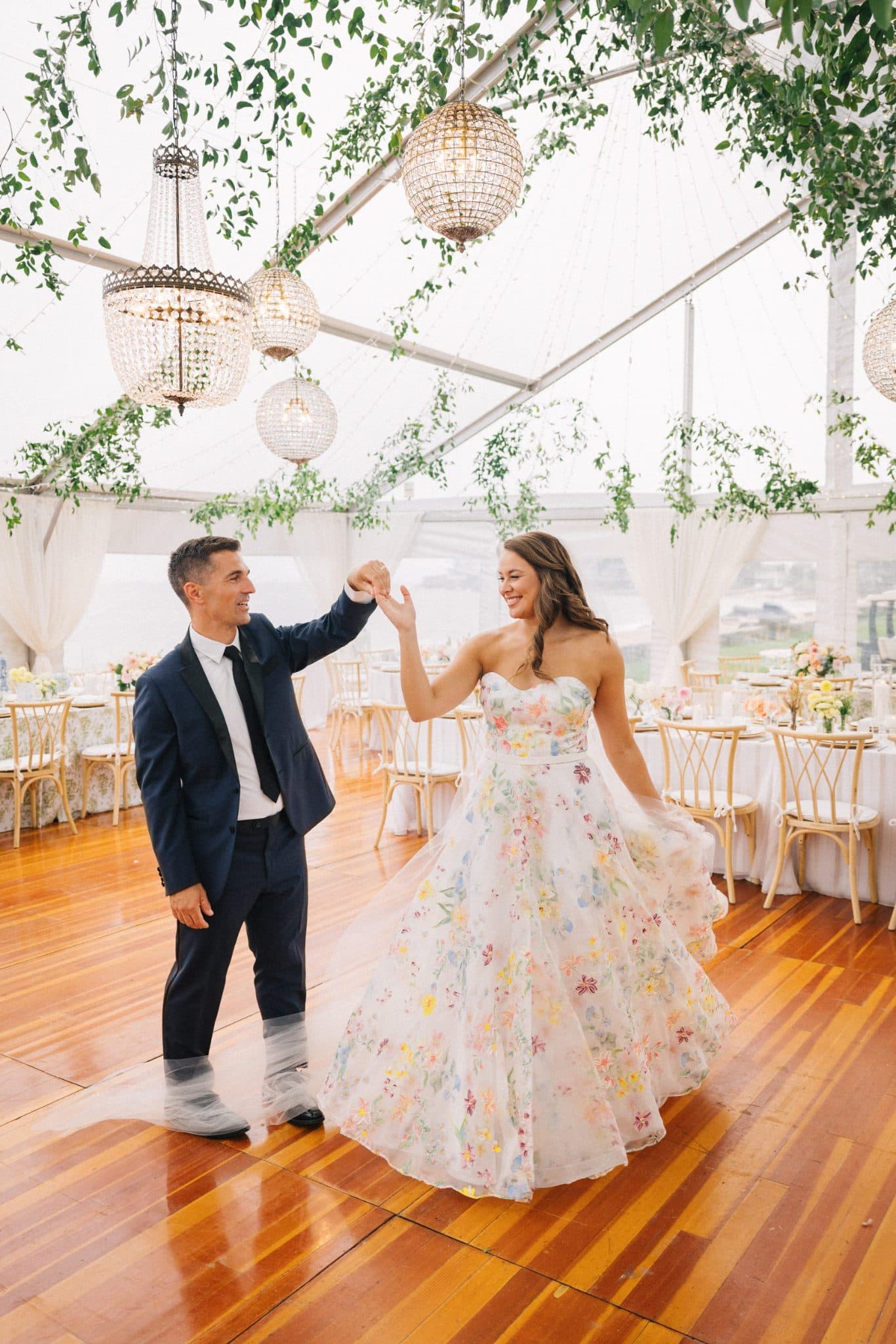 A couple dances joyfully in a tent adorned with hanging lights and greenery.