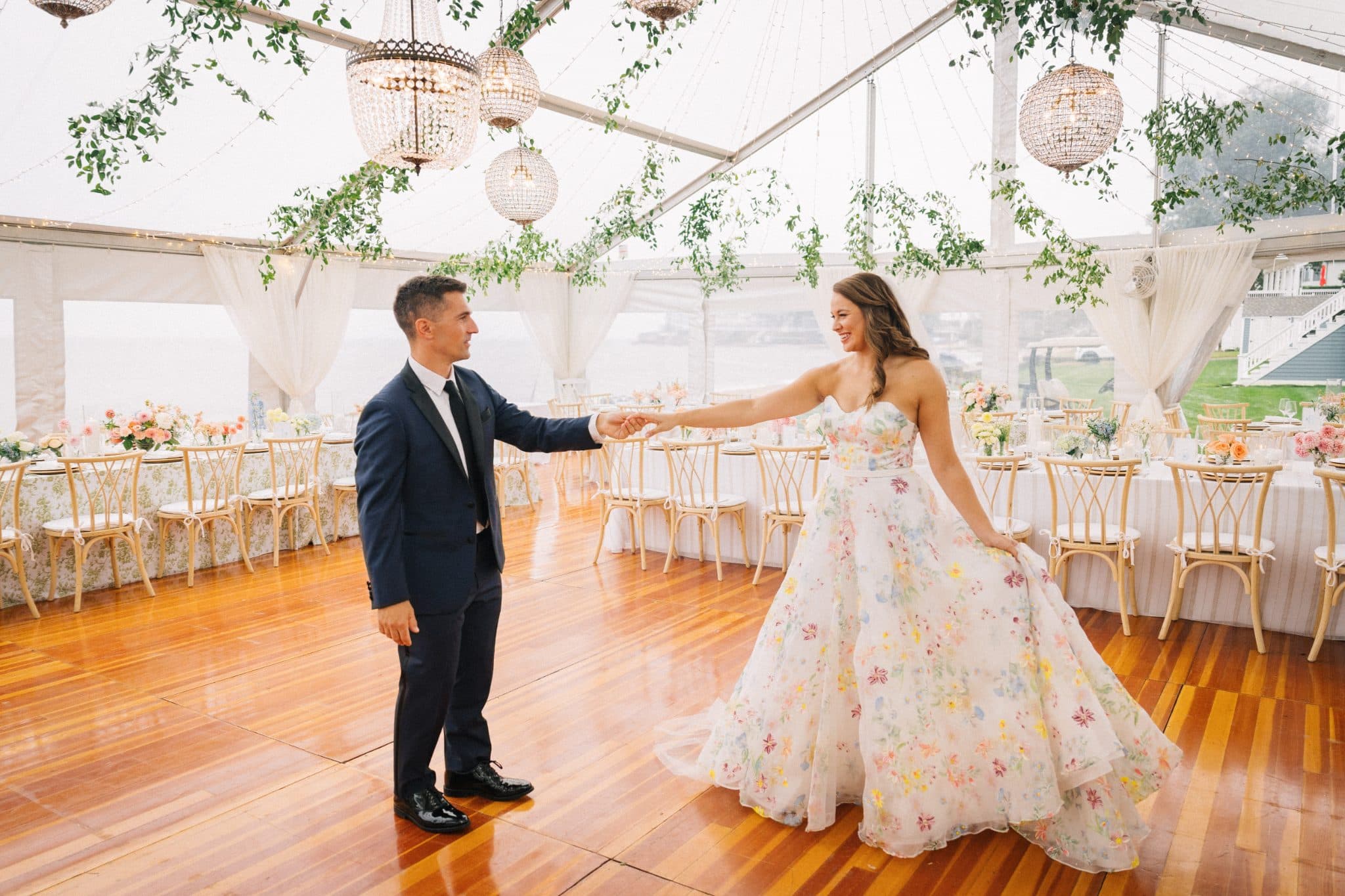 A couple dances in a tent adorned with chandeliers and floral arrangements.