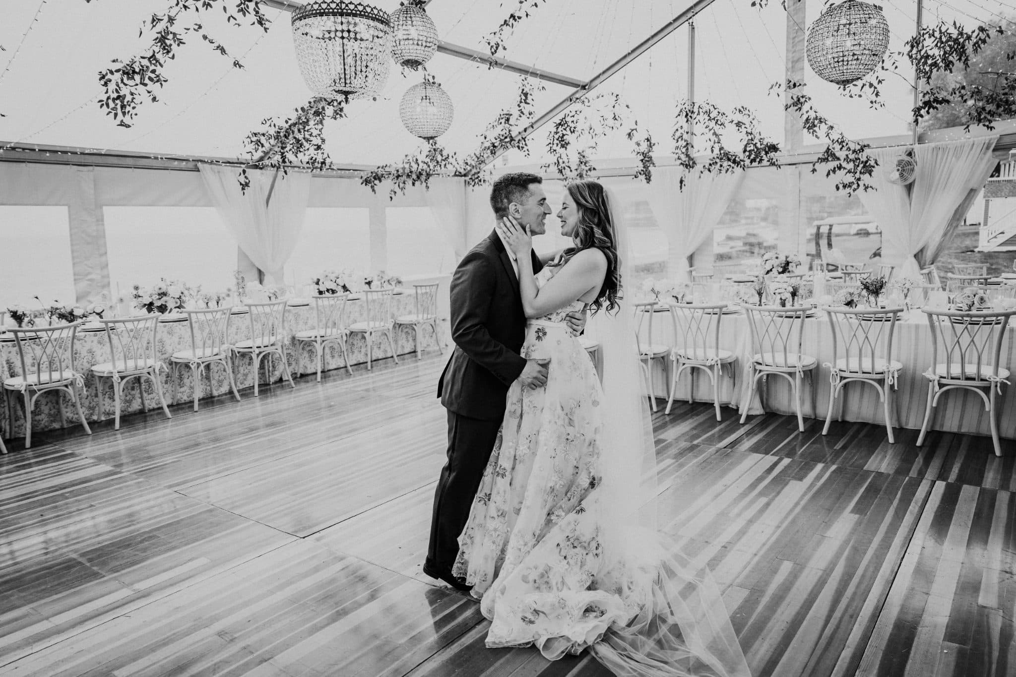 A bride and groom stand close, embracing on a wooden dance floor under a tent with hanging lights and floral decorations.