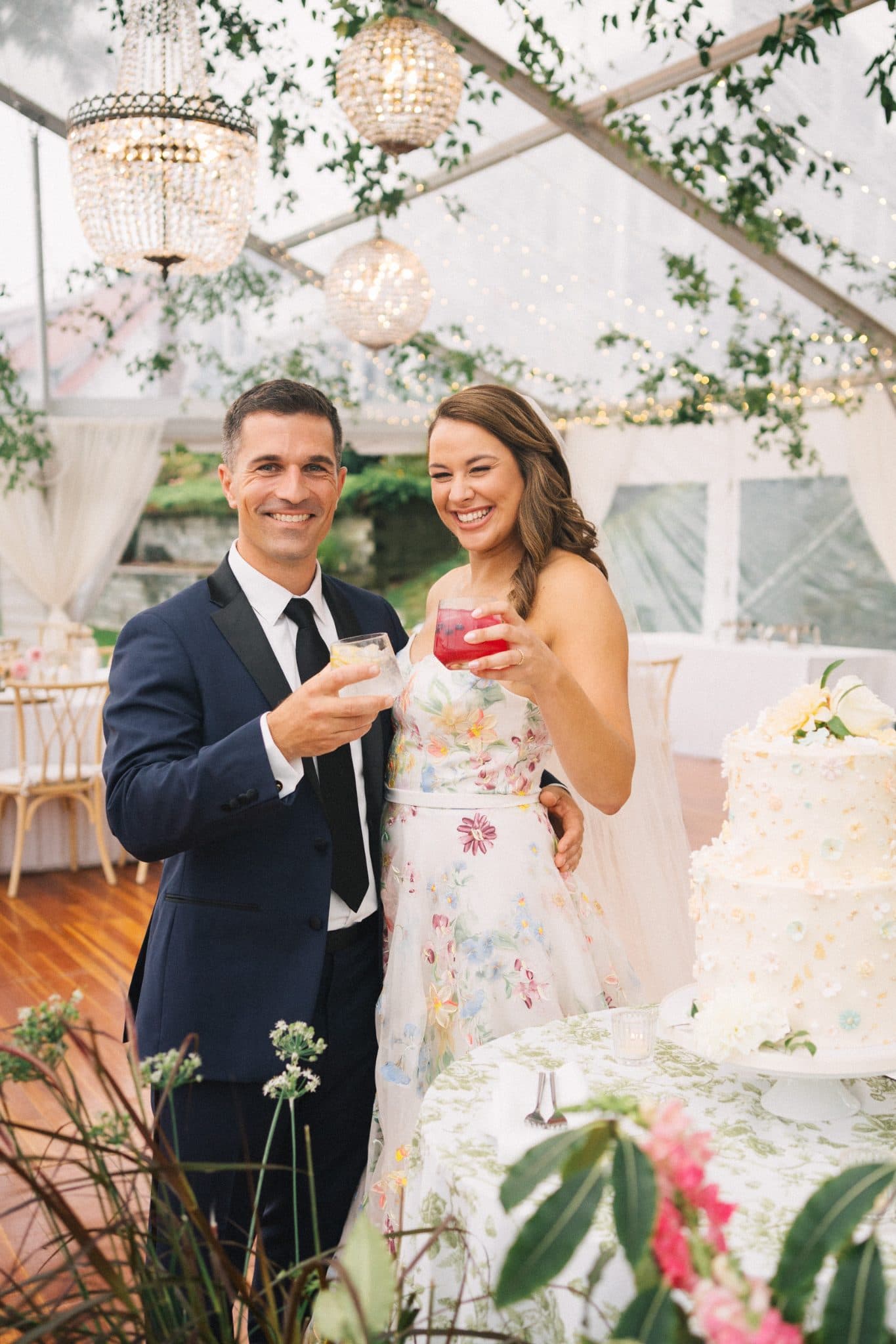 A couple stands together, holding drinks, in front of a floral cake and a decorated tent.