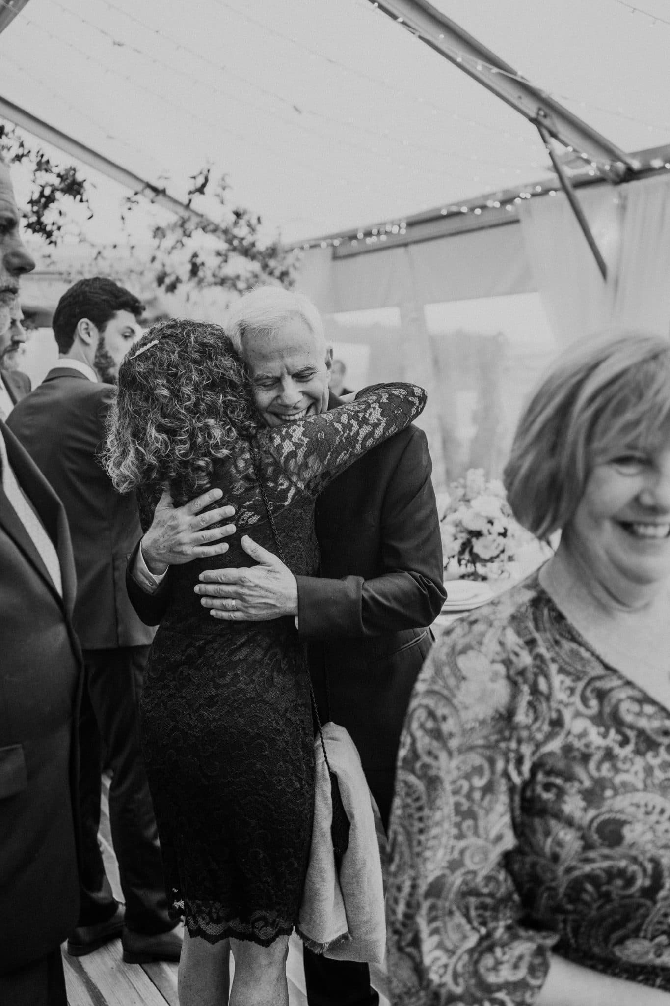 A man in a suit hugs a woman in a lace dress under a tent with string lights.