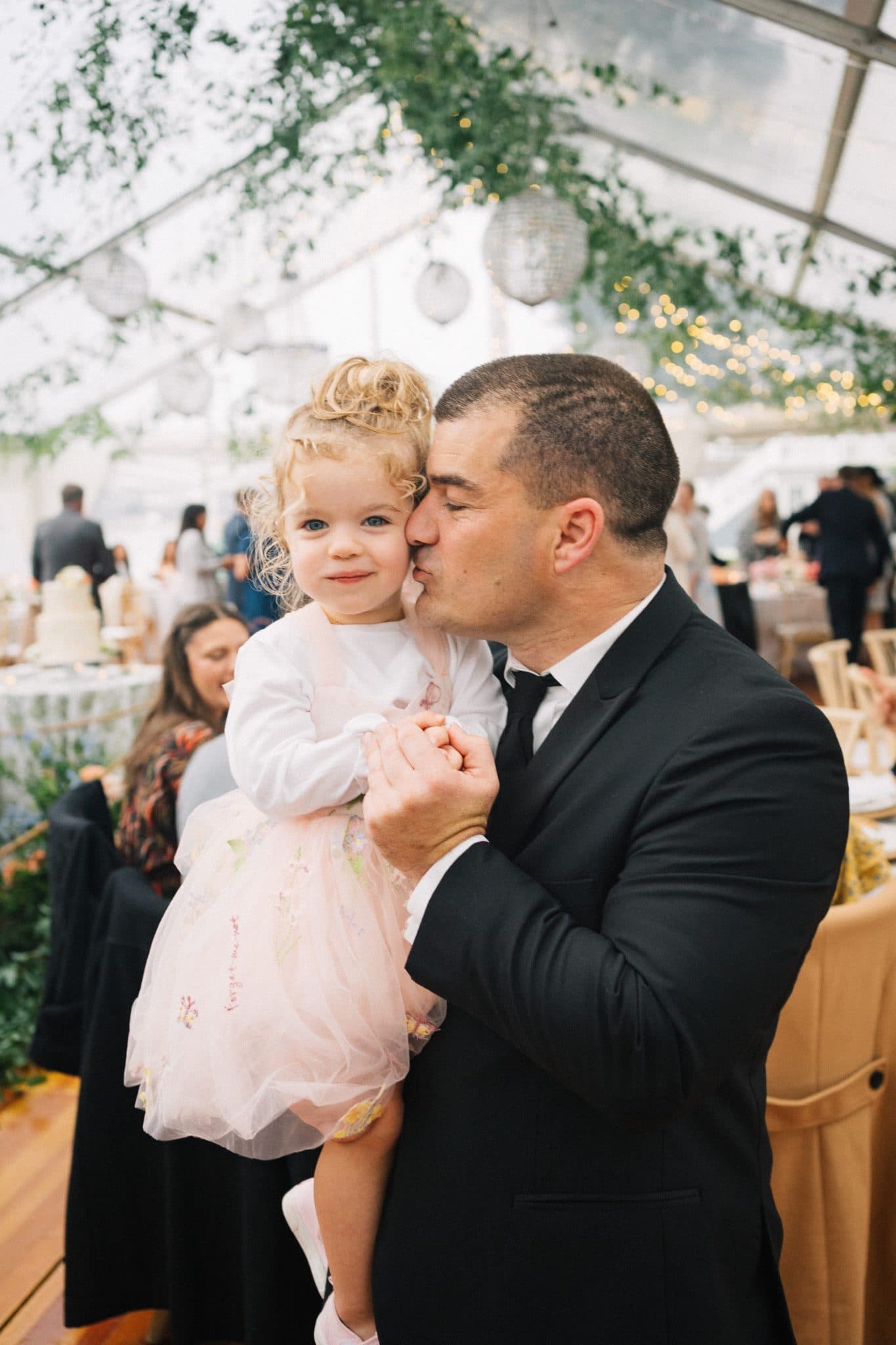 A man in a suit holds a young girl in a pink dress, both smiling, at a wedding reception with guests and decorations in the background.