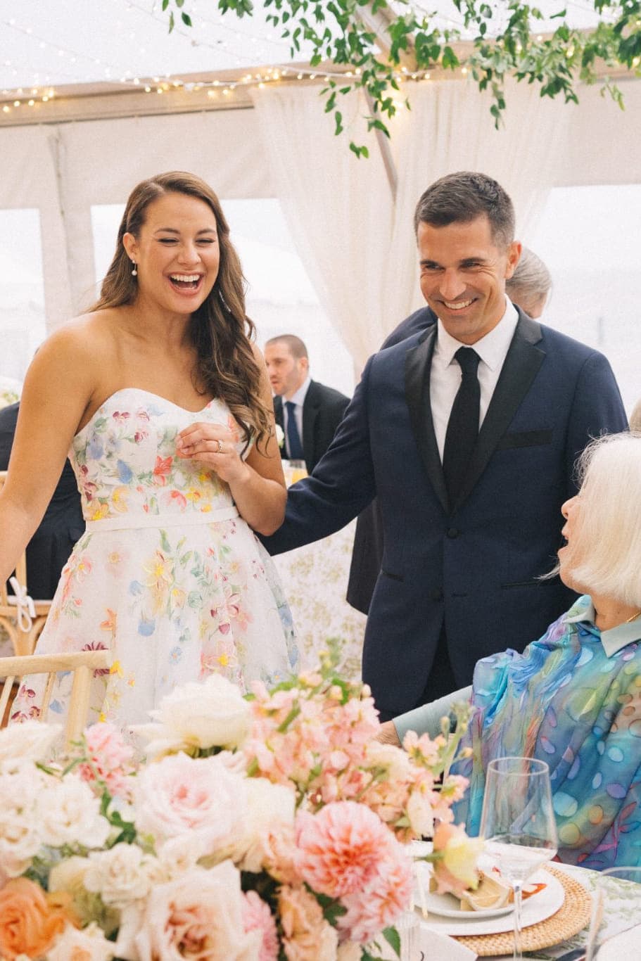 A bride in a floral dress and a man in a suit smile at a seated woman in a colorful dress at a wedding reception.