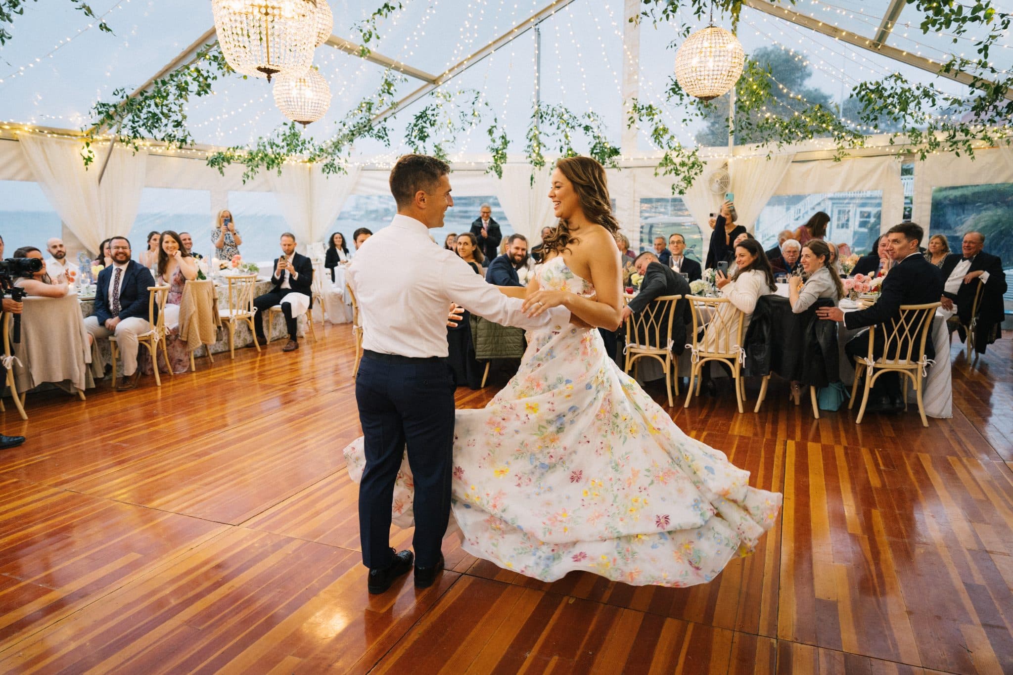 A couple dances joyfully on a wooden floor under a tent adorned with lights and greenery.